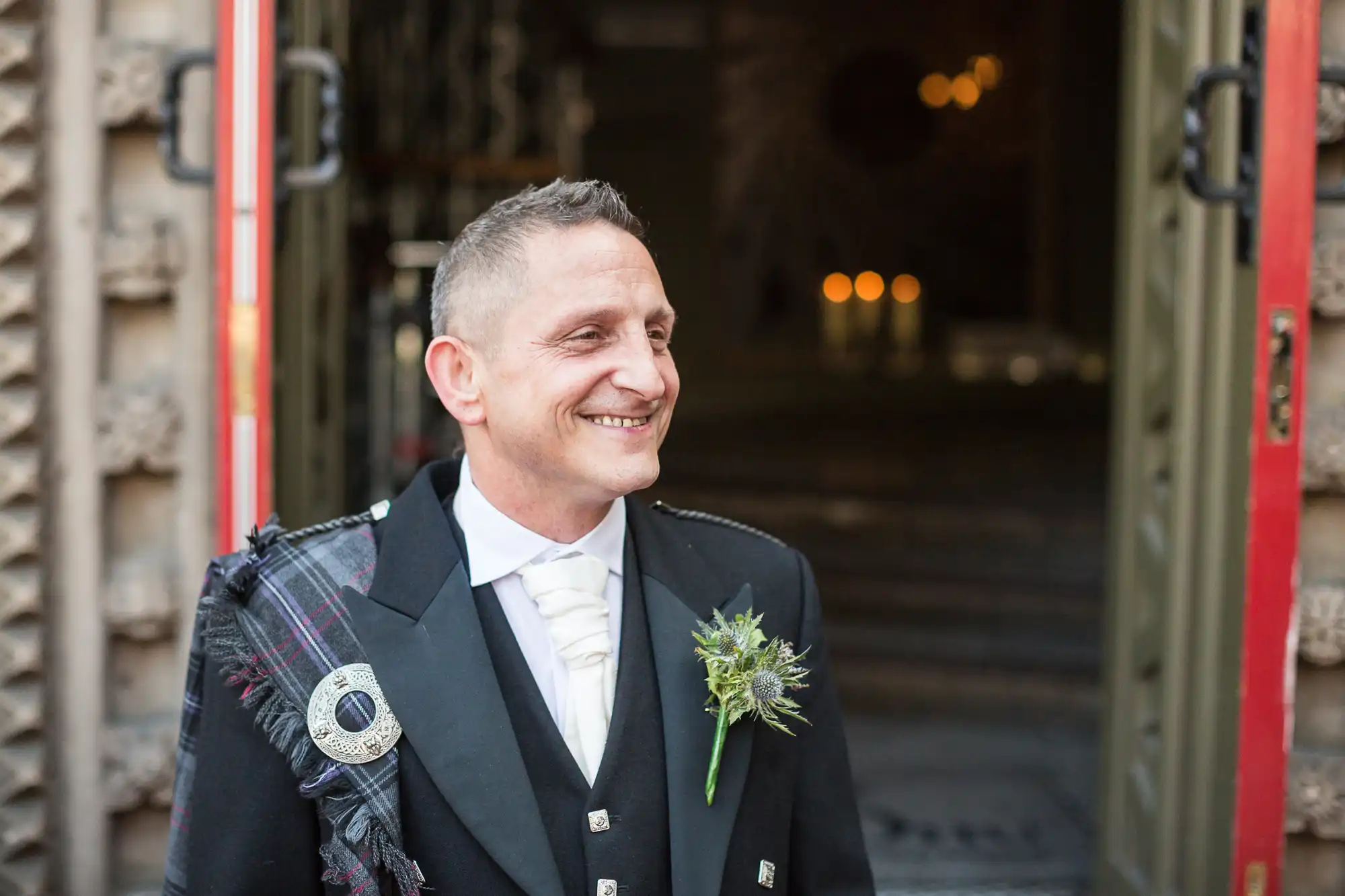 A smiling man in a scottish kilt and jacket with a boutonniere, standing in front of open ornate doors.