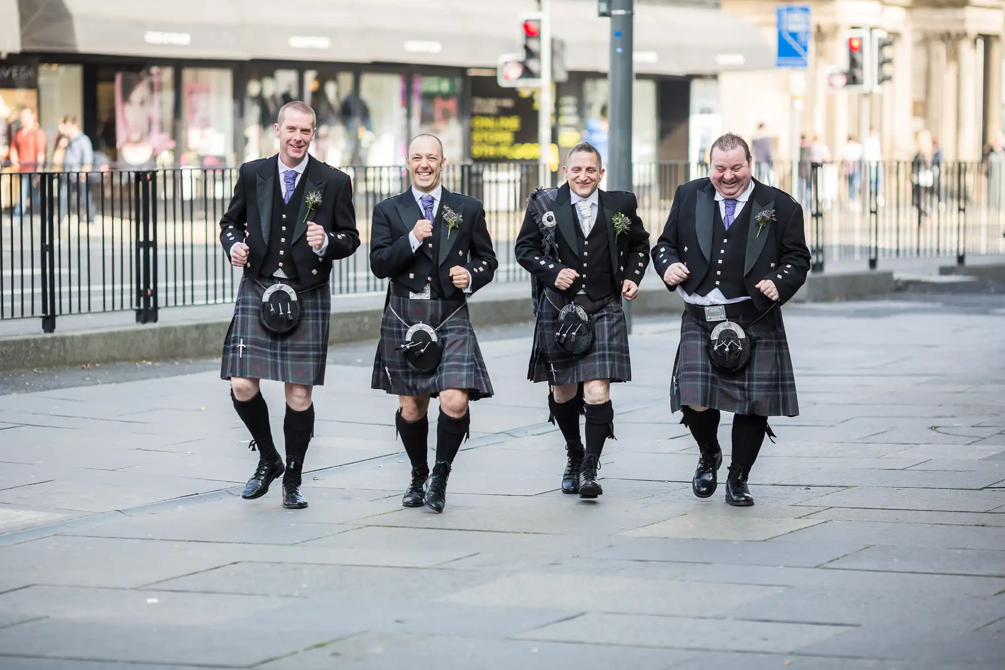 Four men in traditional scottish kilts and jackets, laughing and walking together on a city street.
