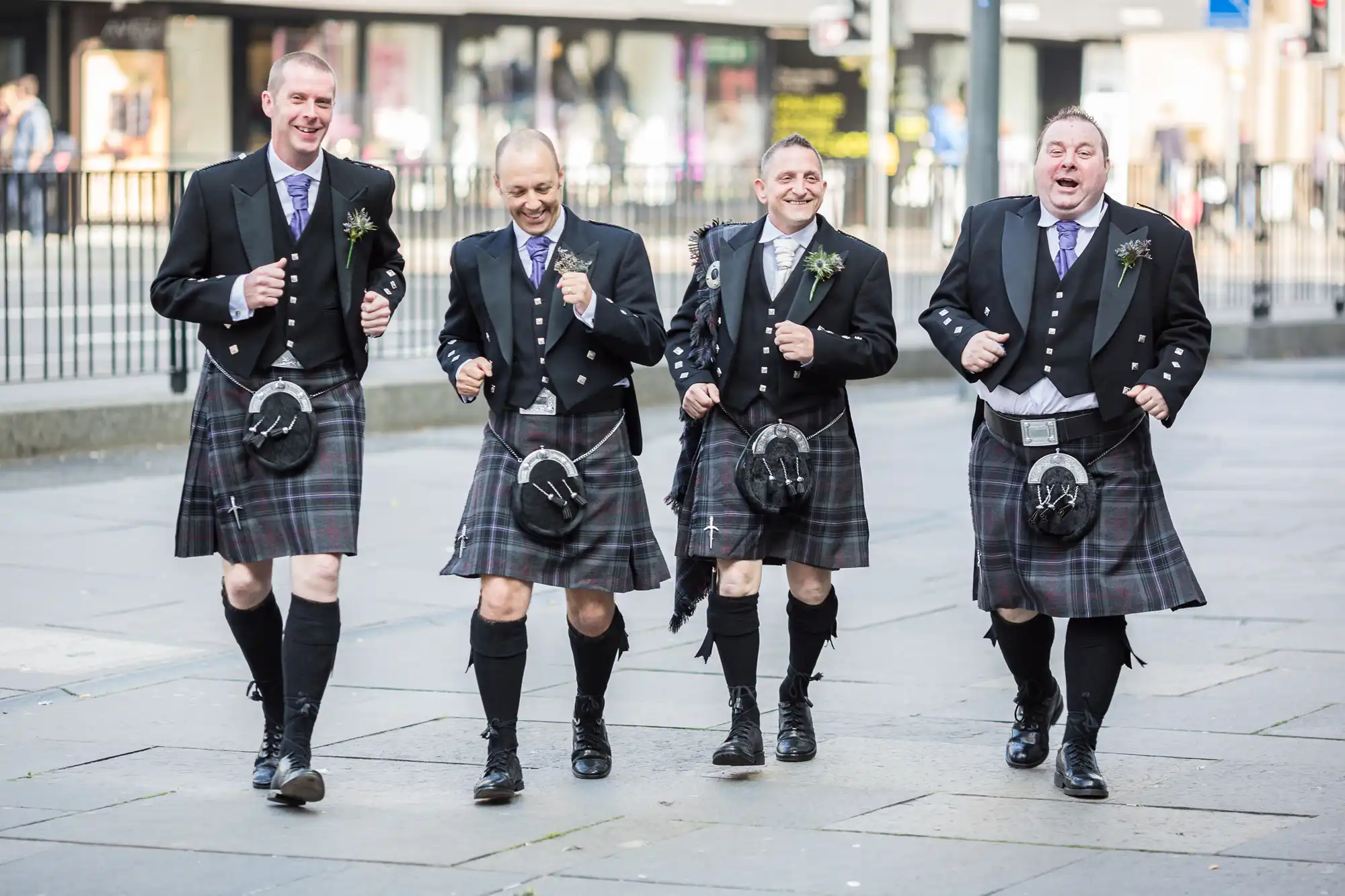 Four men in traditional scottish kilts and jackets joyfully walking down a city street, laughing and holding boutonnières.