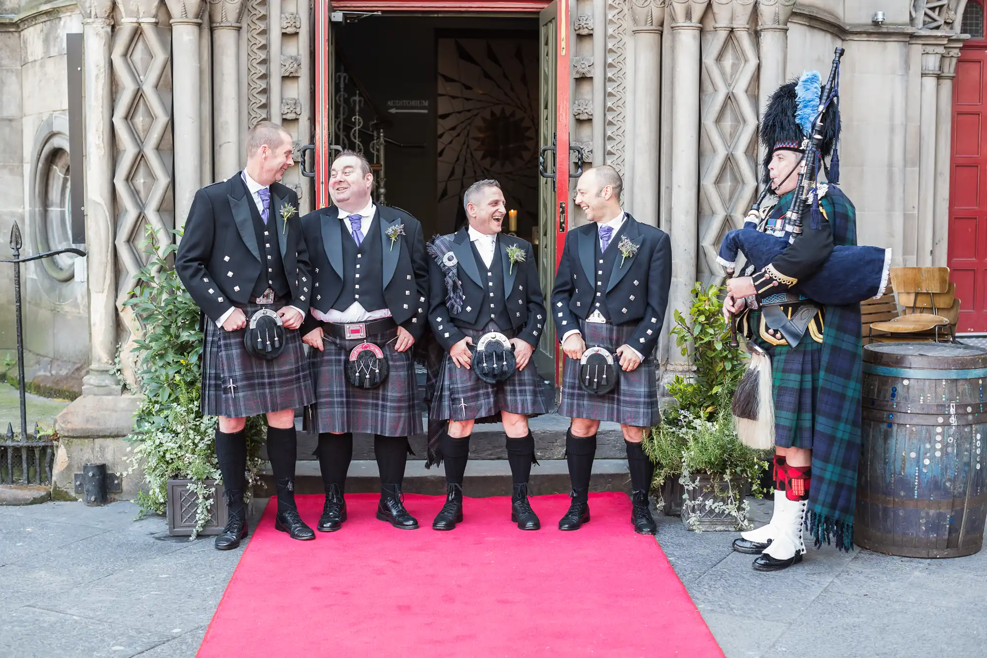 Four men in traditional scottish kilts laugh together outside a building, while a piper in full highland dress plays nearby.