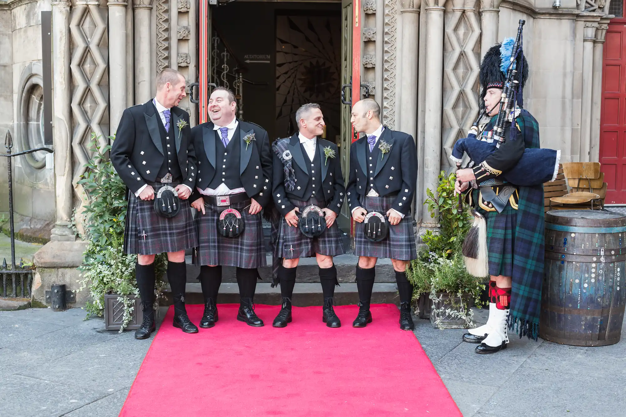 Five men in traditional scottish kilts laughing together outside a building entrance, with a bagpiper in full attire standing to the side.