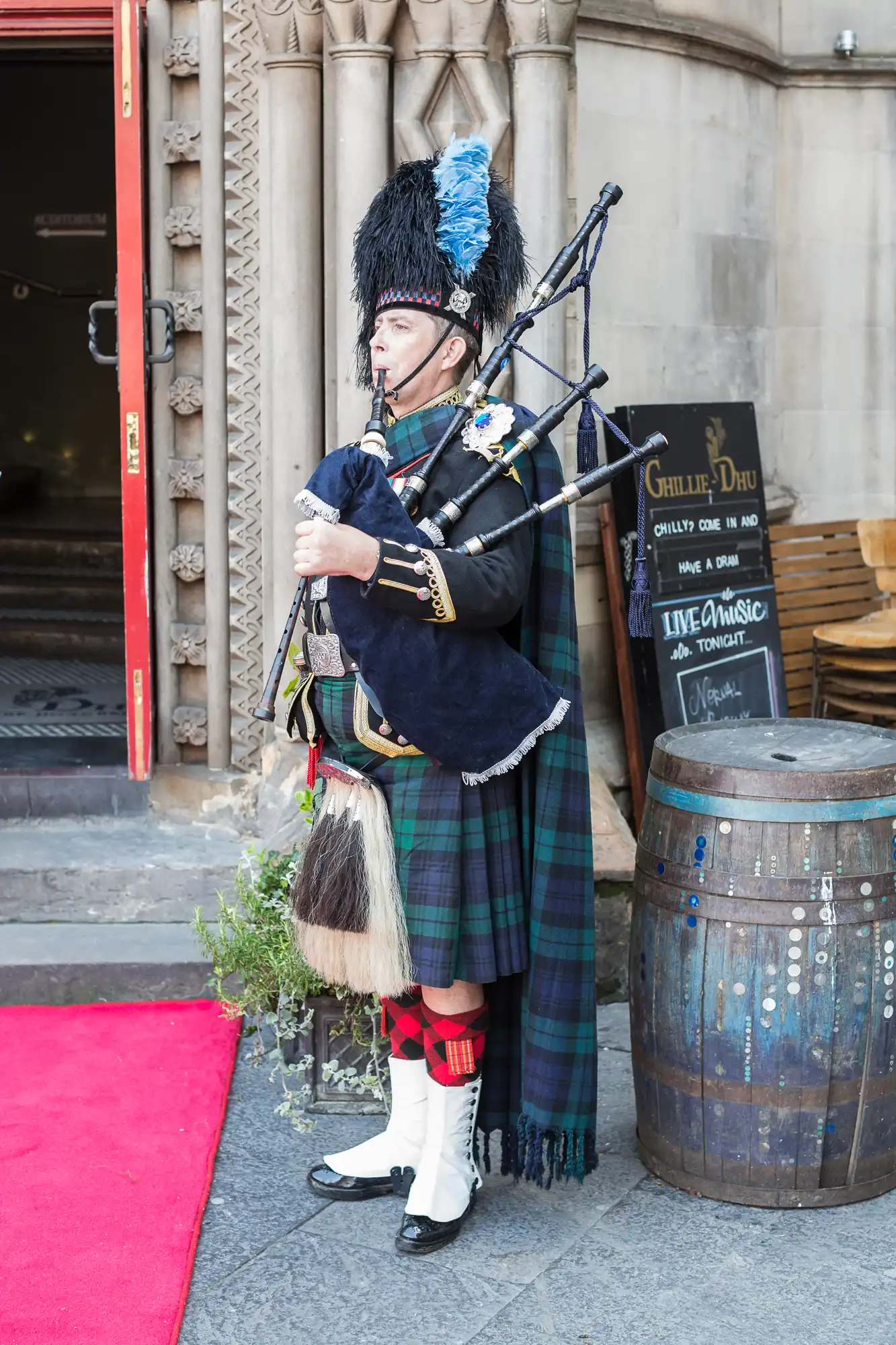 A man in traditional scottish attire playing bagpipes outside a building with a sign promoting live music.
