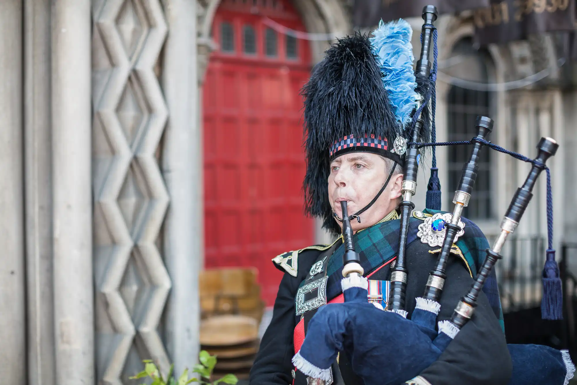 A man in traditional scottish attire playing bagpipes in front of a red door.