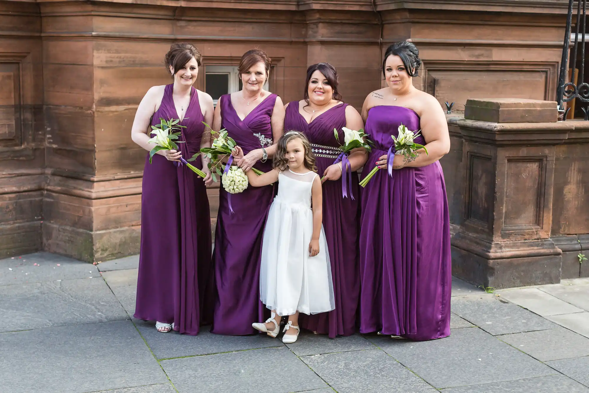 Four women in purple dresses holding bouquets and a young girl in a white dress smiling, standing together outside a brick building.
