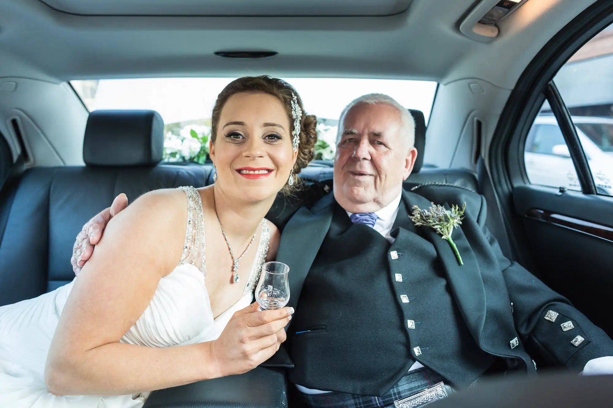 A bride in a white dress sits next to an older man in a kilt and suit inside a car, both smiling at the camera, the bride holding a champagne glass.