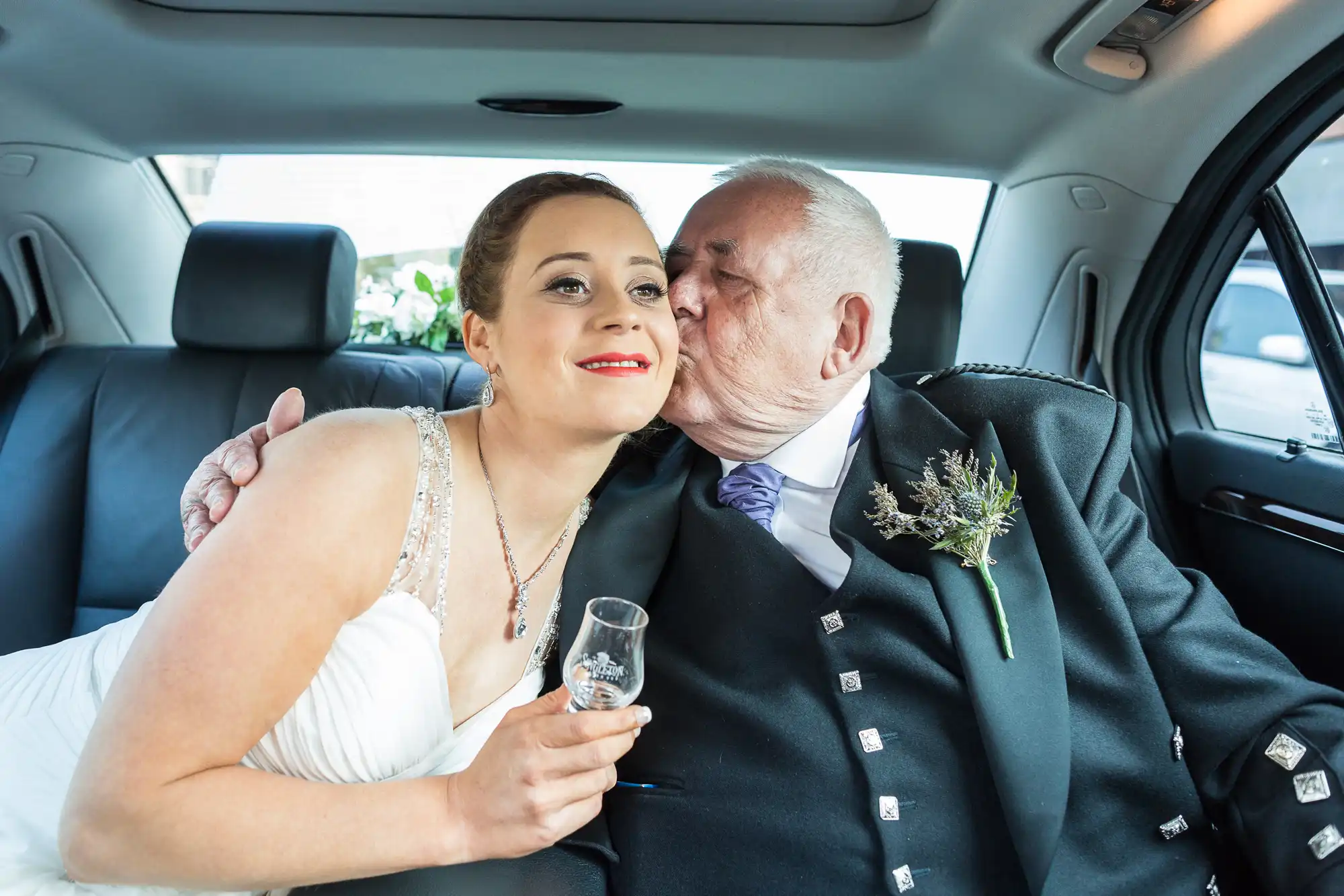 A bride in a white dress receives a kiss on the cheek from an older man in a suit inside a car.