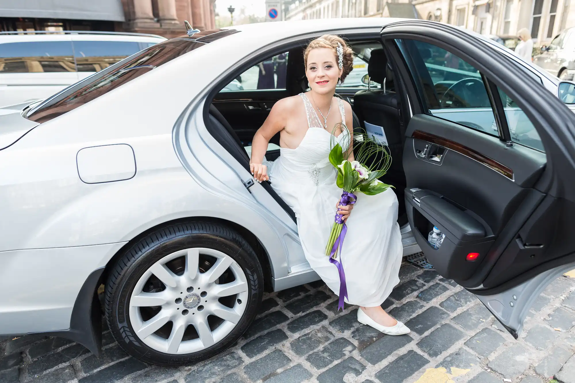 A bride in a white dress and pearl necklace steps out of a silver car holding a bouquet of green and purple flowers.