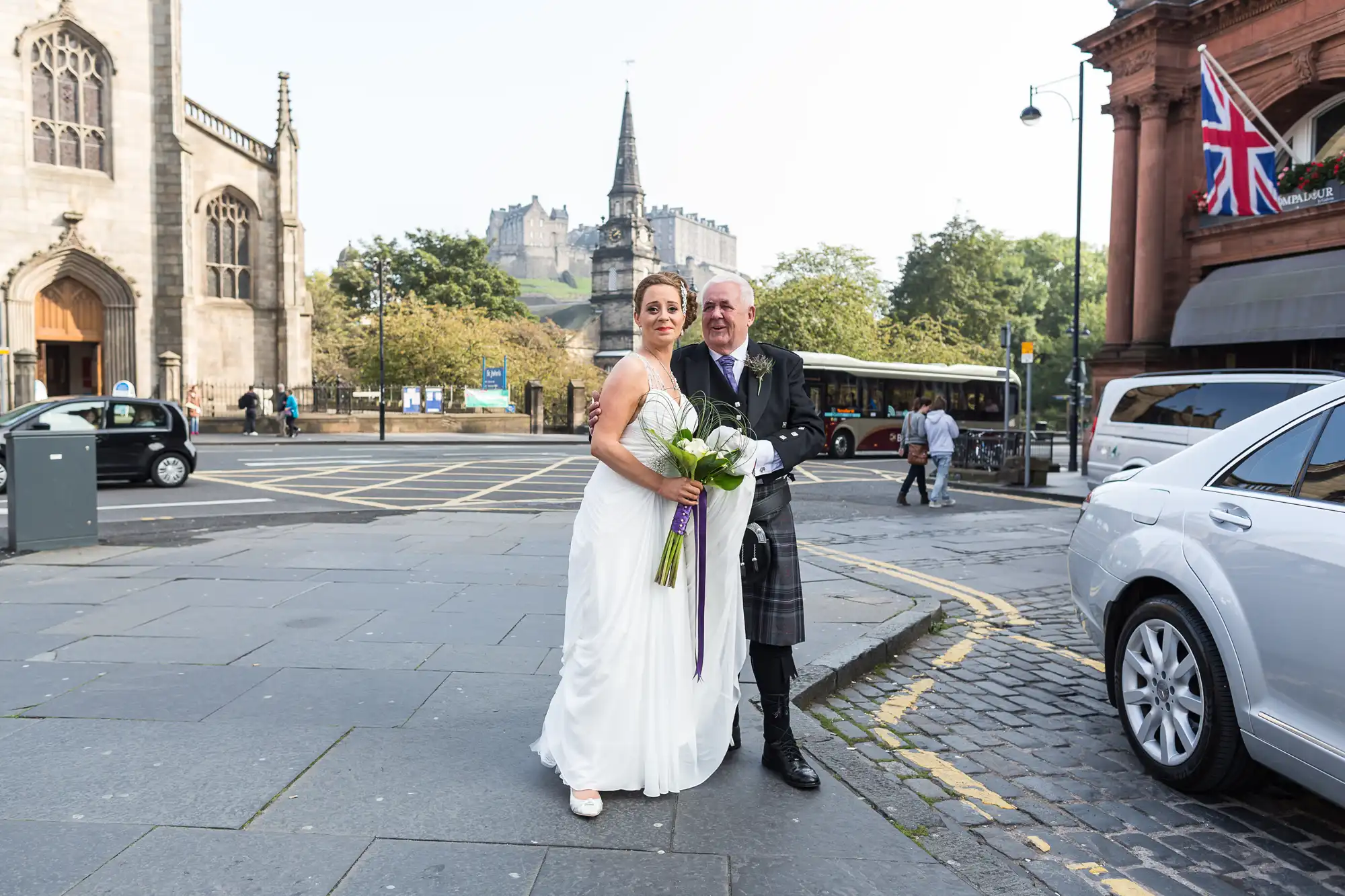 A bride in a white dress and a groom in a kilt stand on a city street, with historic buildings and a british flag in the background.
