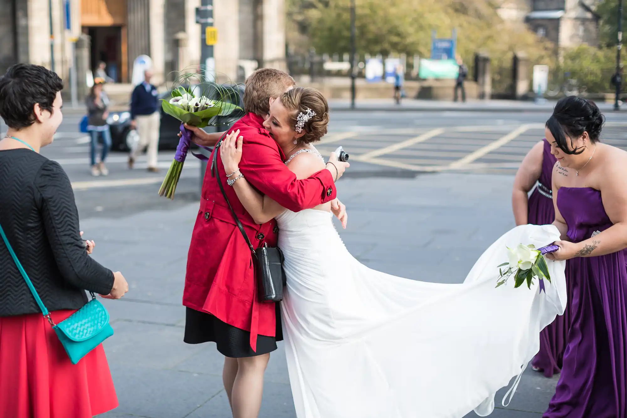 A bride and groom embrace on a city street, surrounded by guests in colorful attire, as one bridesmaid holds the bride's long train.
