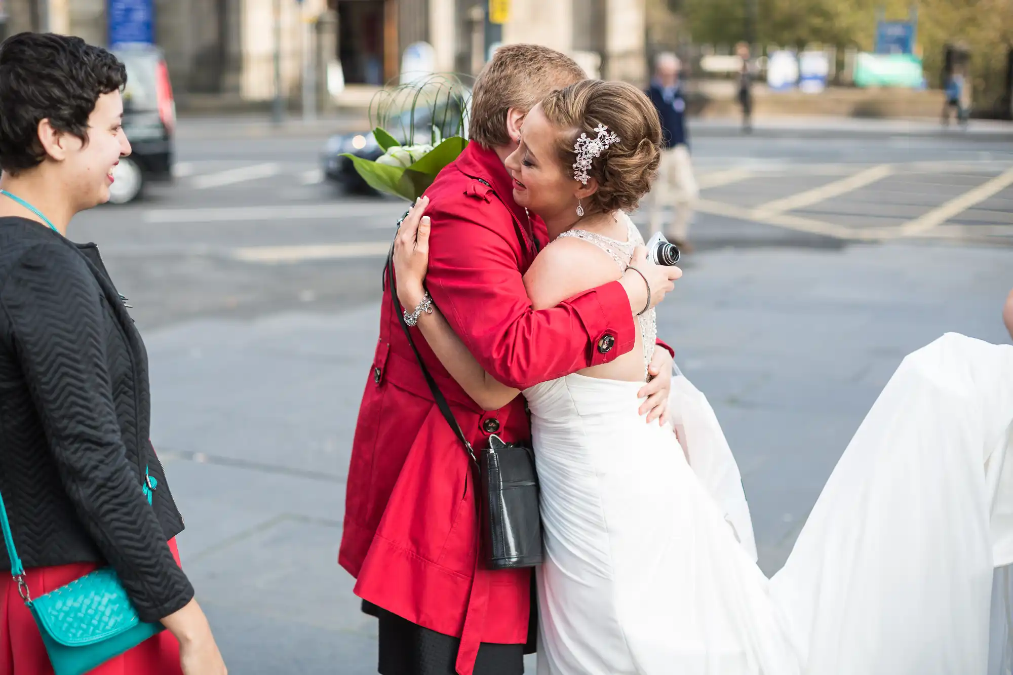 A bride in a white dress embracing a woman in a red jacket on a city street, with bystanders watching.
