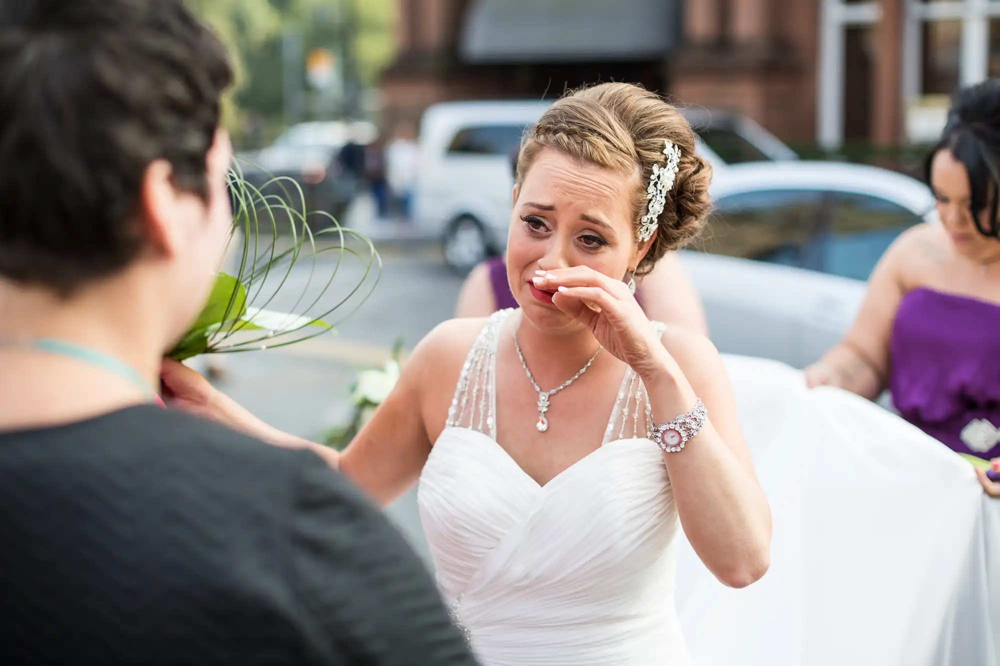 A bride in a white gown wiping tears from her eyes while holding a bouquet, speaking to a woman in a dark dress at an outdoor wedding.