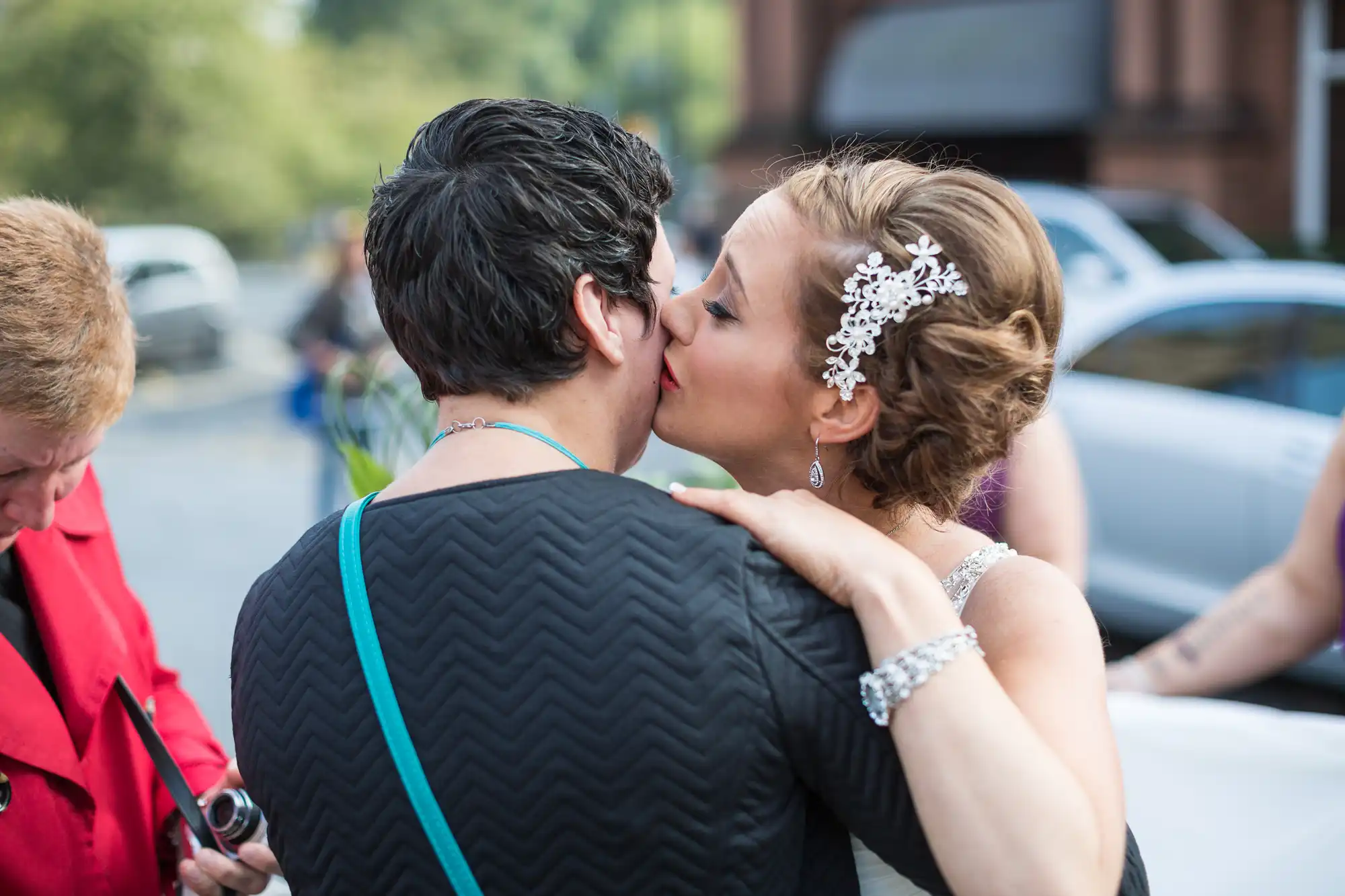 A bride and groom kissing, the bride with a decorative hairpiece, on a street with a woman and cars in the background.