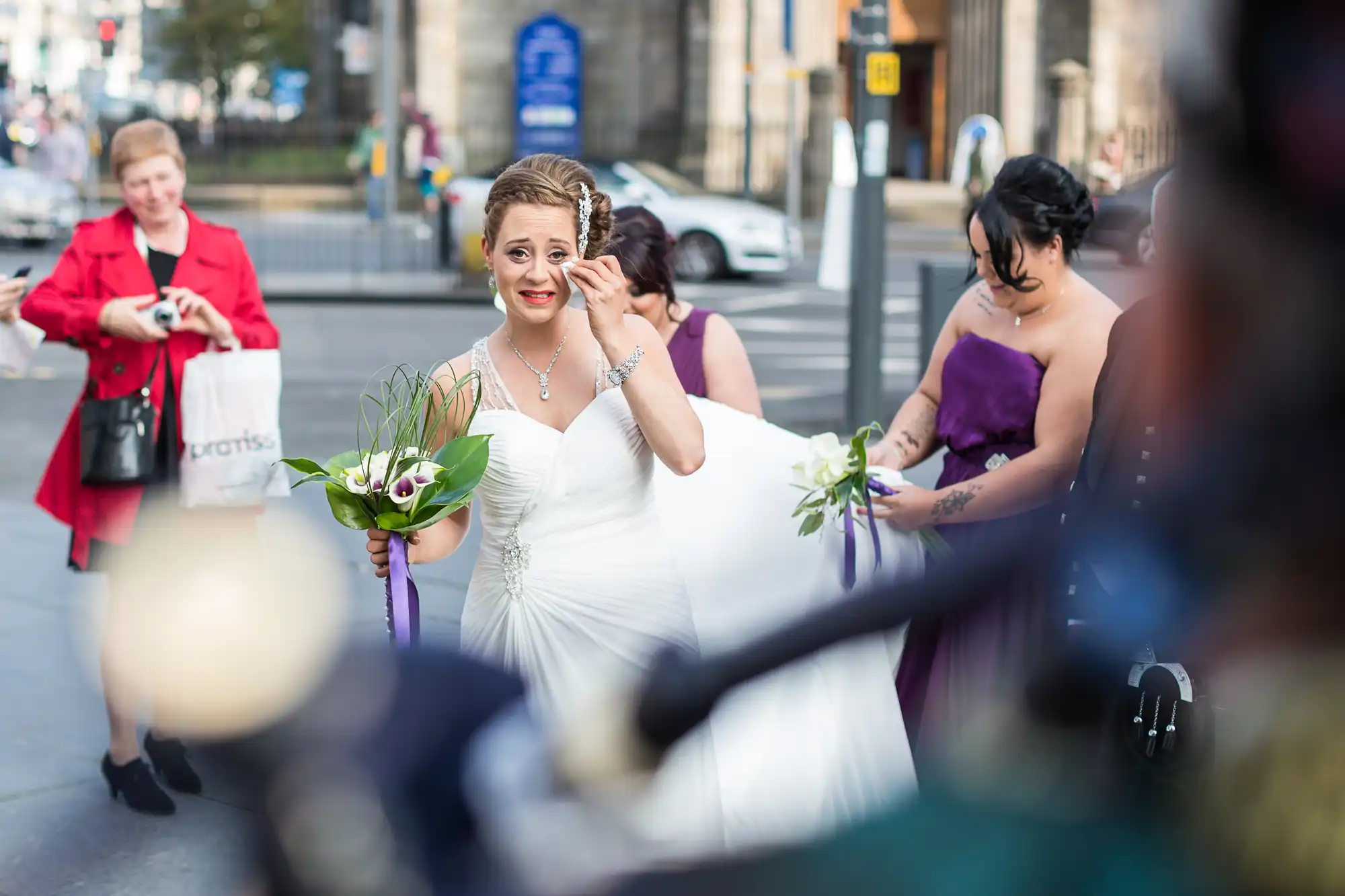 A bride holding a bouquet looks surprised on a busy city street, with guests in colorful attire and a photographer capturing the moment.