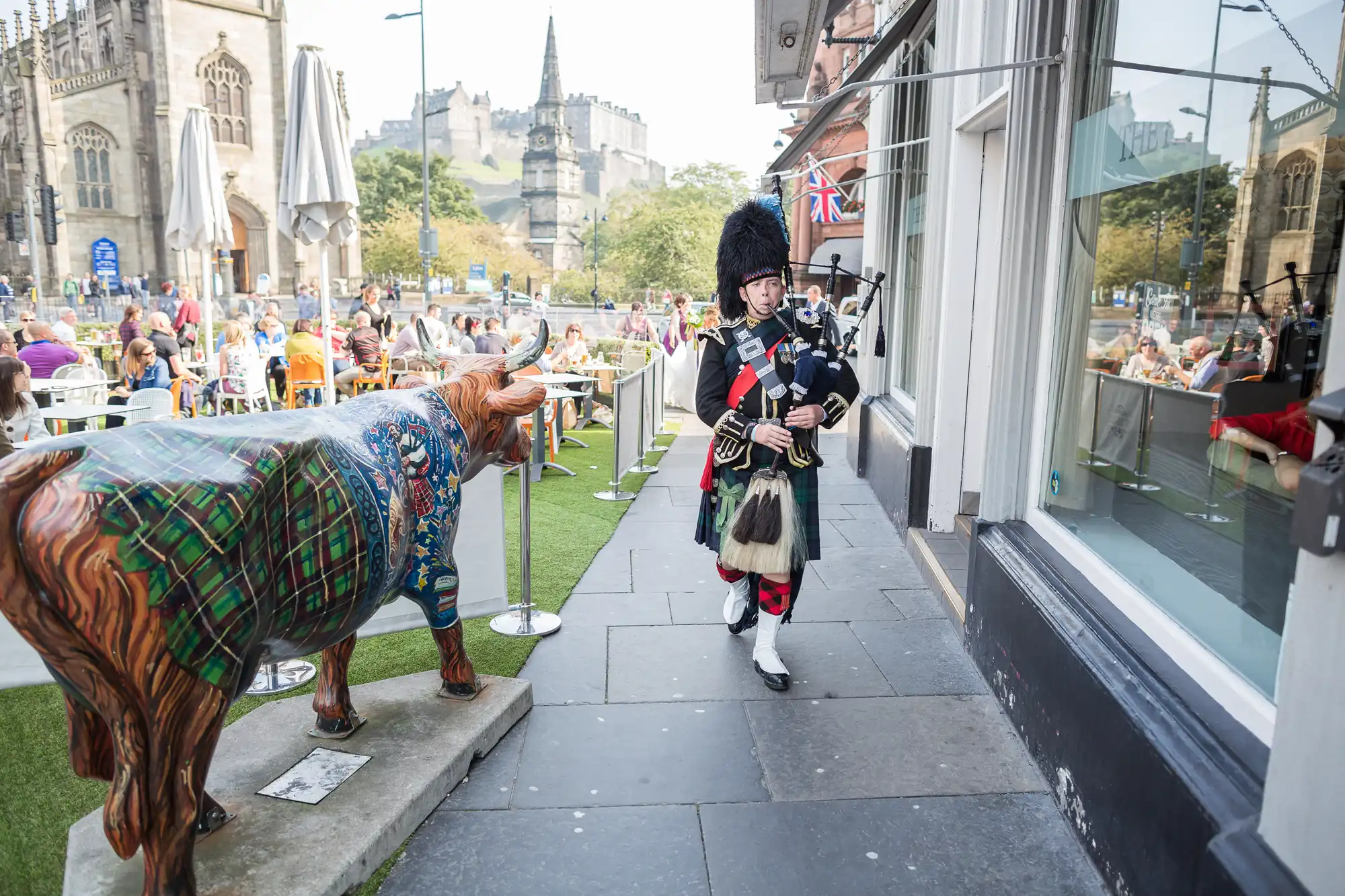 A bagpiper in traditional scottish attire walks by a busy outdoor cafe and tartan-painted cow statues in an urban setting.