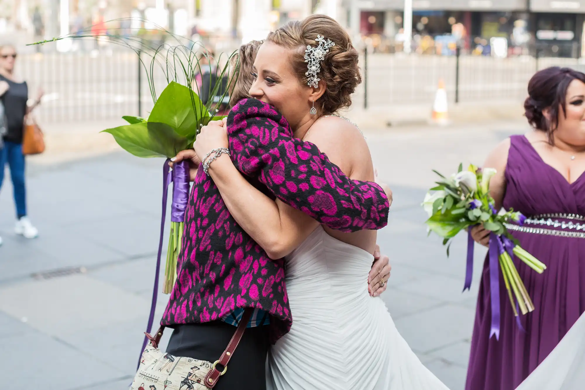 A bride in a white dress embracing a woman in a pink leopard print jacket on a city street, with a bridesmaid in purple nearby.