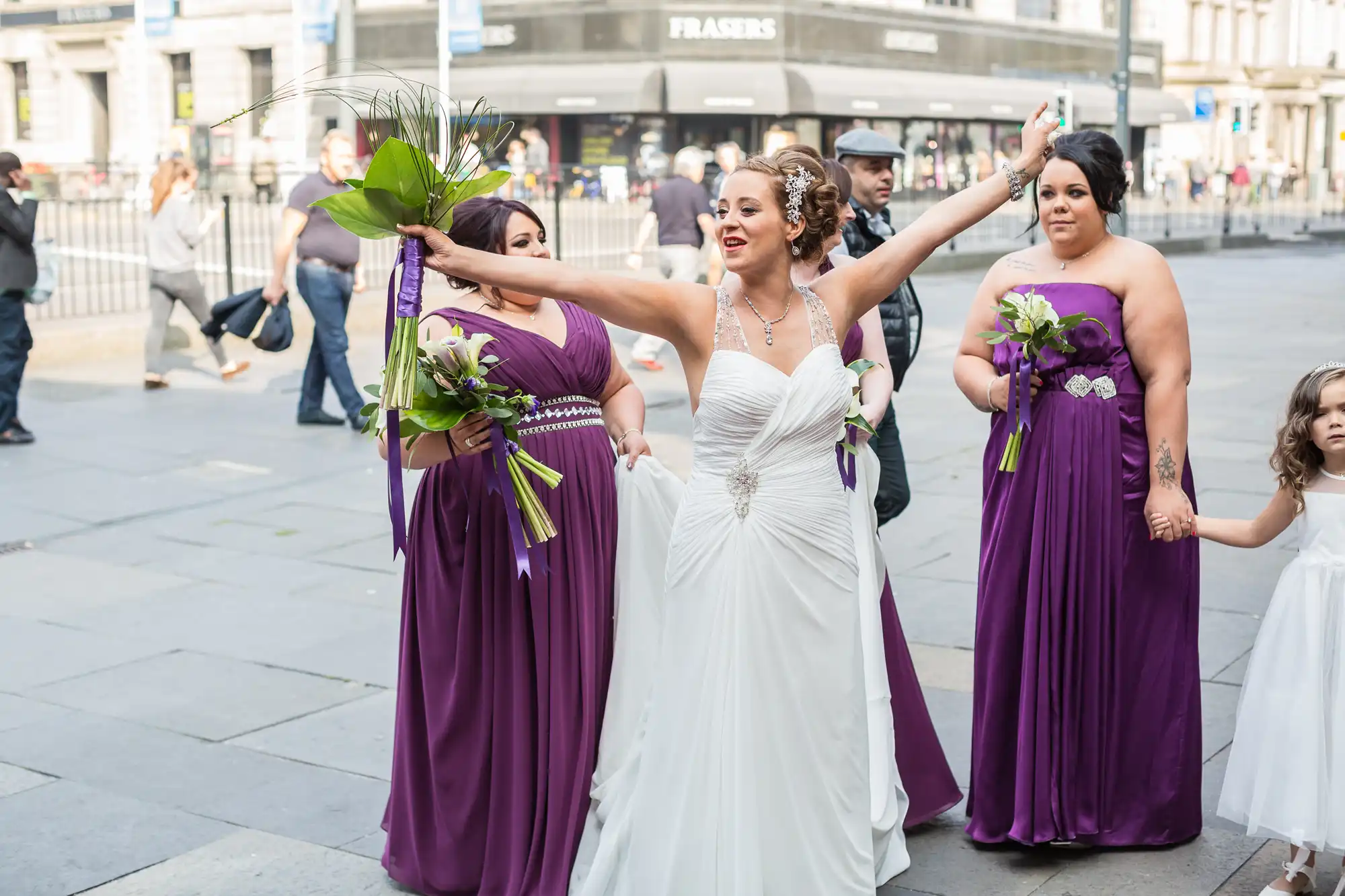 A bride in a white dress and three bridesmaids in purple dresses walking through a busy street, with bystanders in the background.