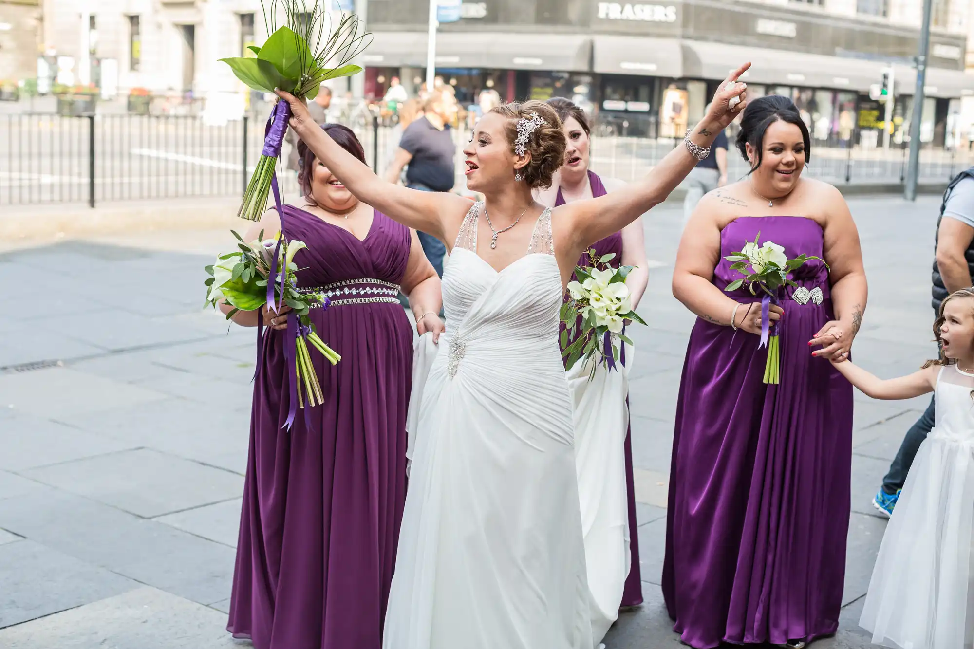 A bride in a white dress and three bridesmaids in purple dresses joyfully walking on a city street, holding bouquets.