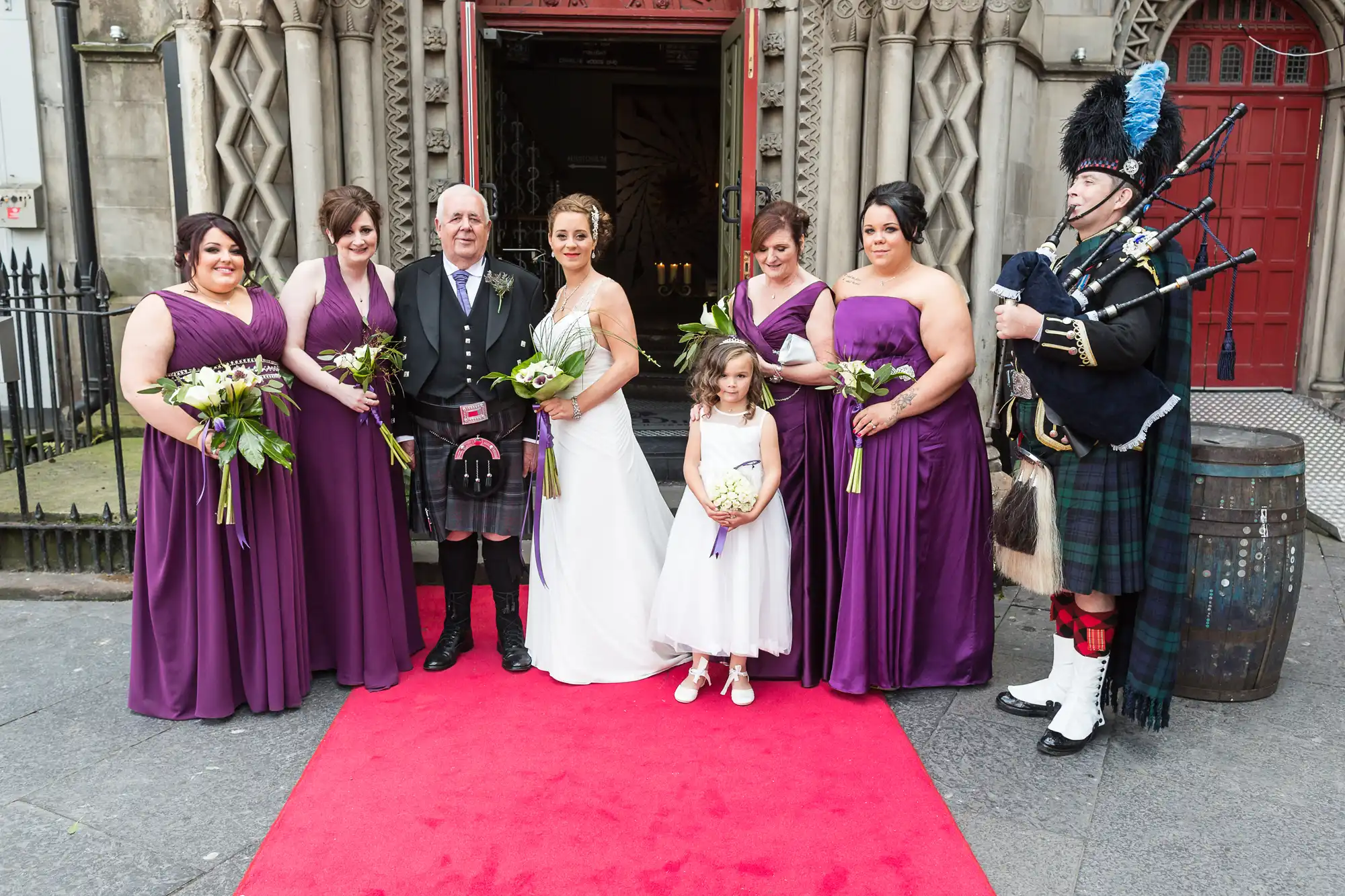 A wedding group posing with a bagpiper outside a church, the women in purple dresses and the men in kilts.