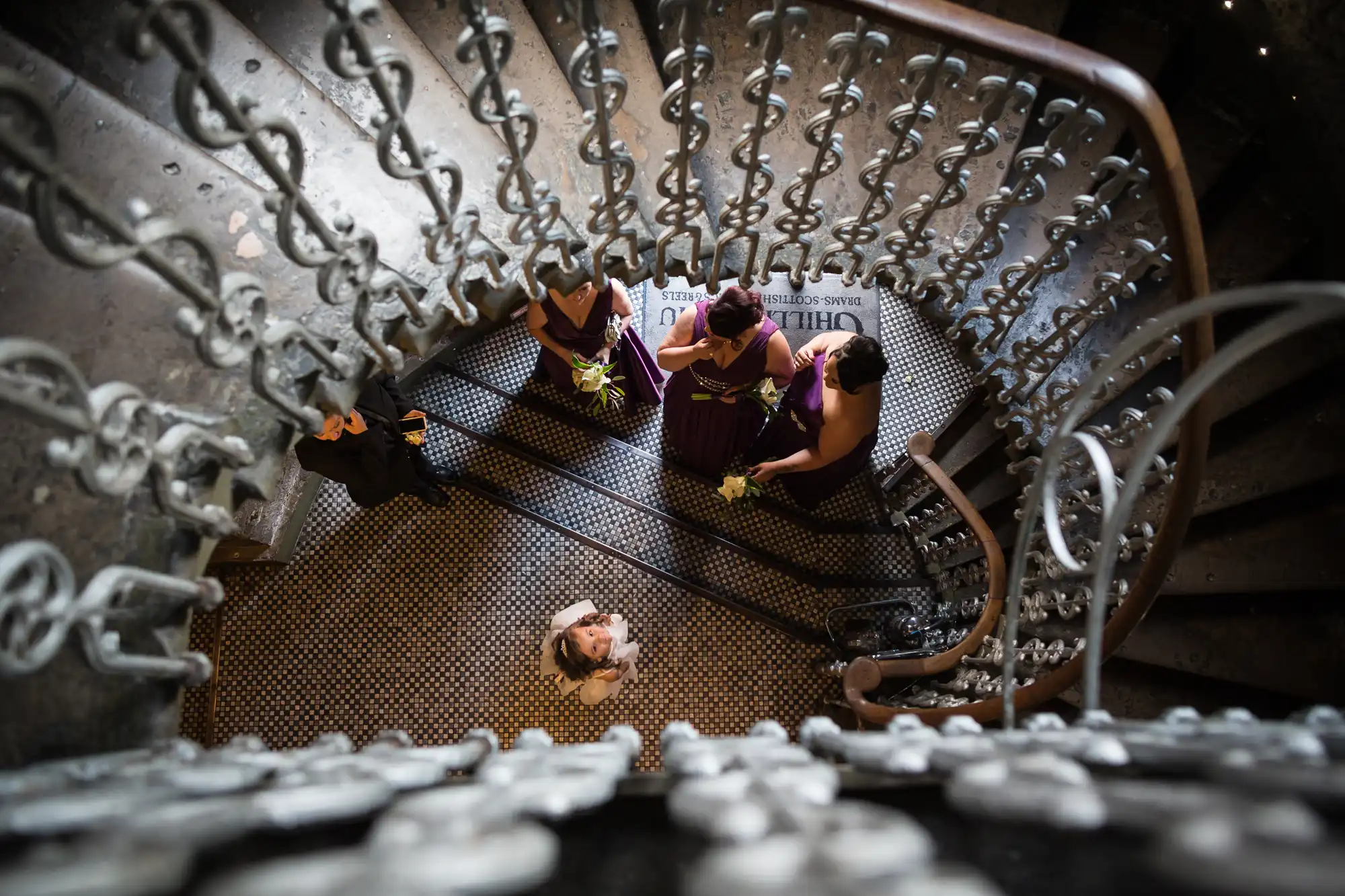 A bride looking up while standing at the bottom of an ornate spiral staircase, surrounded by bridesmaids dressed in purple.