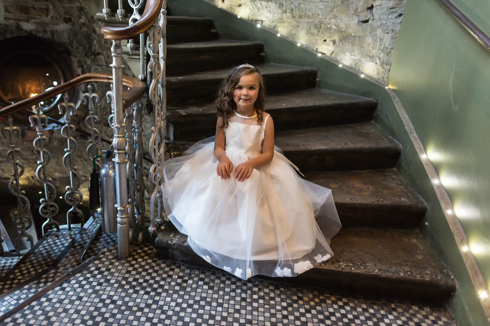 A young girl in a white dress sits smiling on a stone staircase with ornate metal railings, in a dimly lit setting.