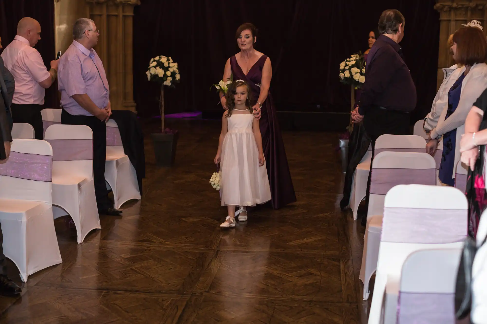 A young girl and a woman in formal dresses walk down an aisle at a wedding ceremony, with guests watching from their seats.