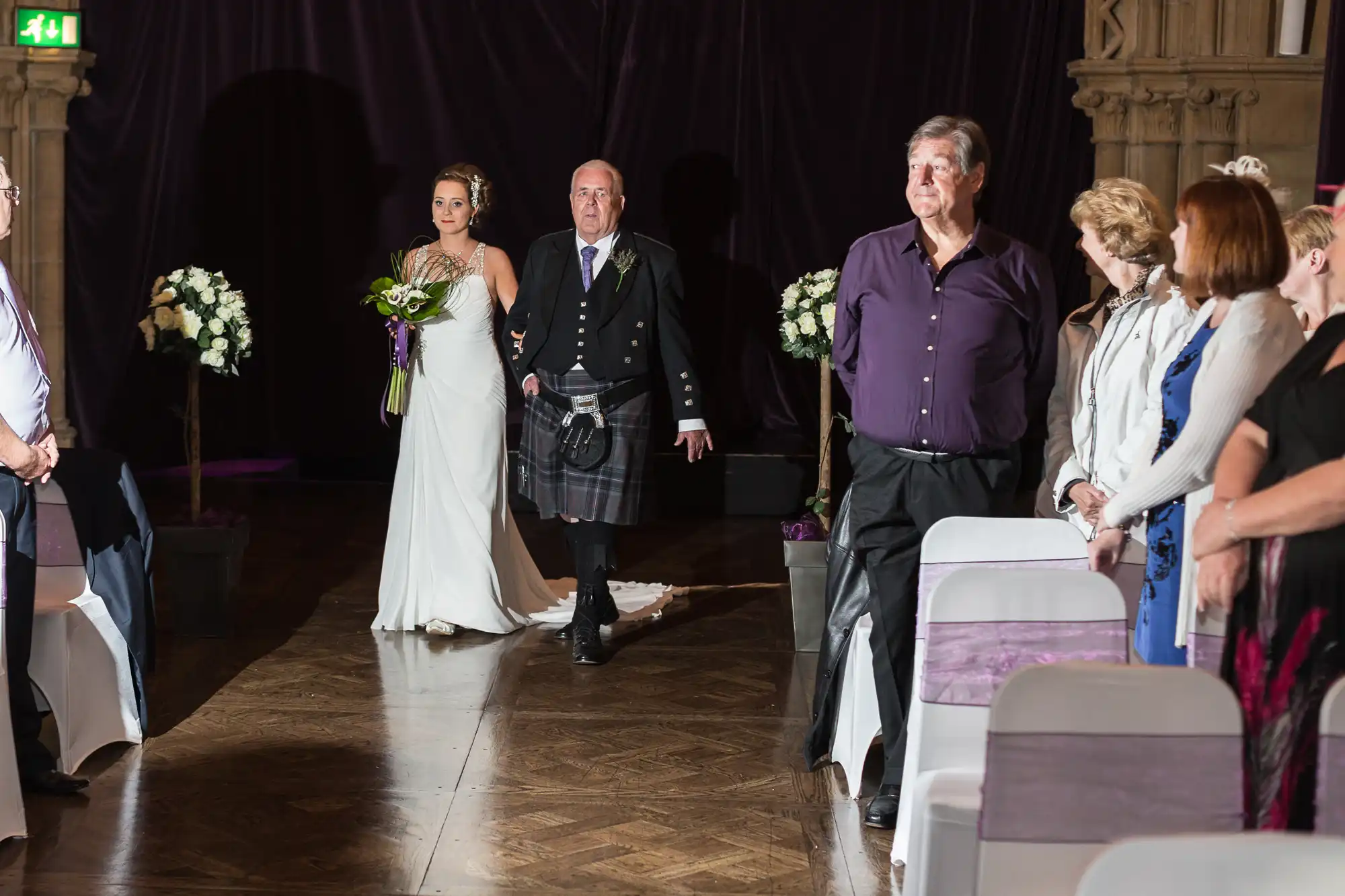 A bride and her father walk down the aisle past seated guests in a dimly lit venue, the father in a kilt and the bride holding a bouquet.