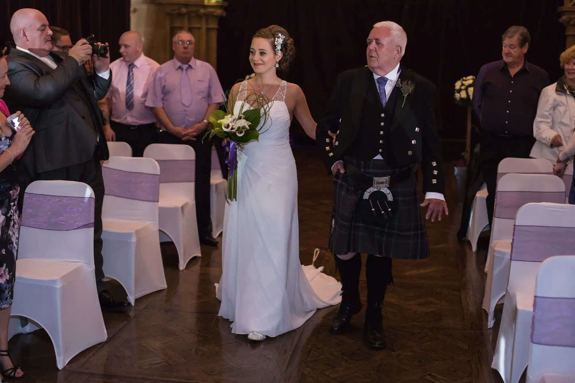 A bride in a white dress walks down the aisle with an older man in a kilt, surrounded by guests capturing the moment with cameras.