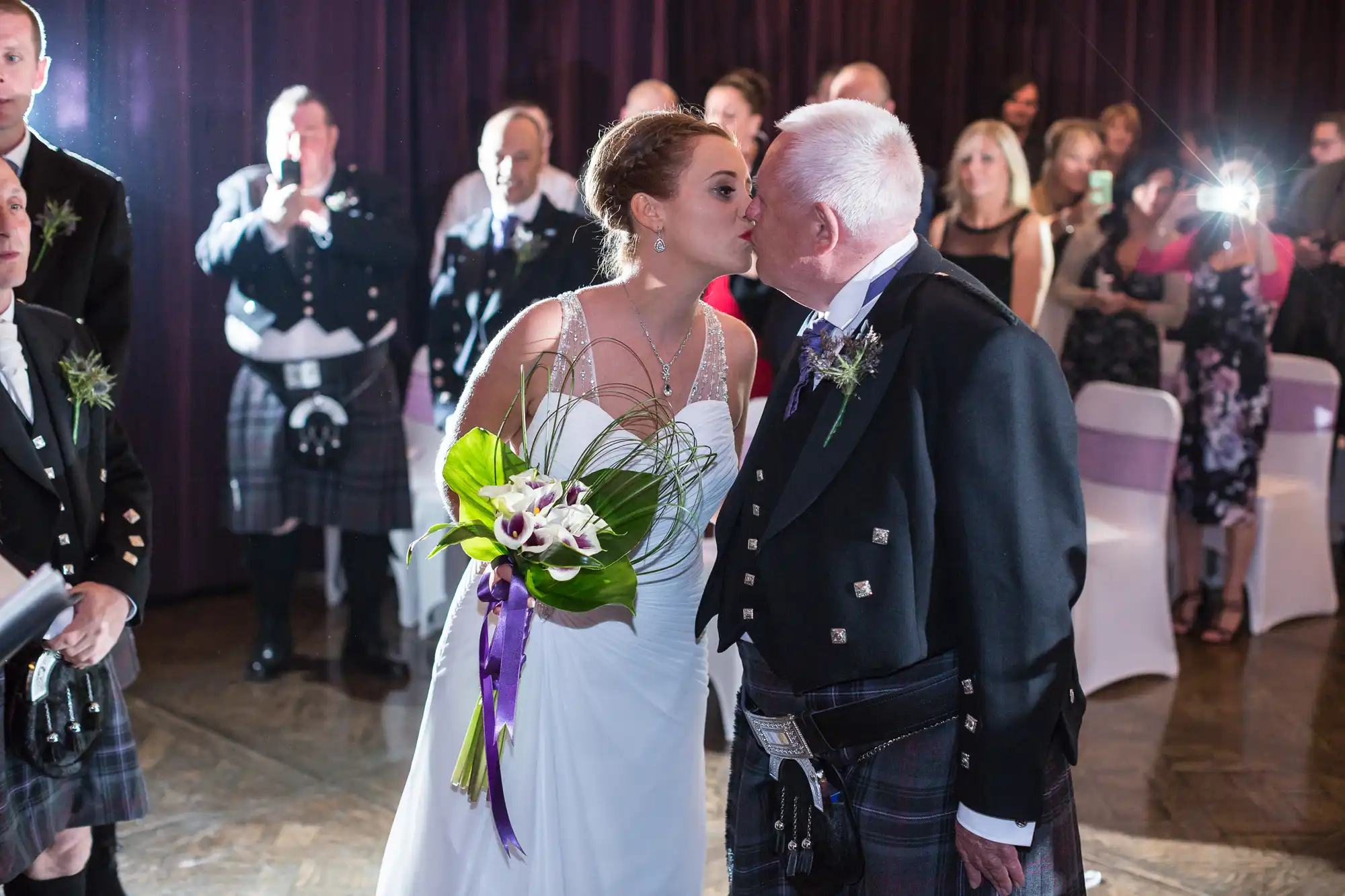 A bride in a white dress kisses an elderly man on the cheek at a wedding reception, surrounded by guests in formal attire, some wearing kilts.