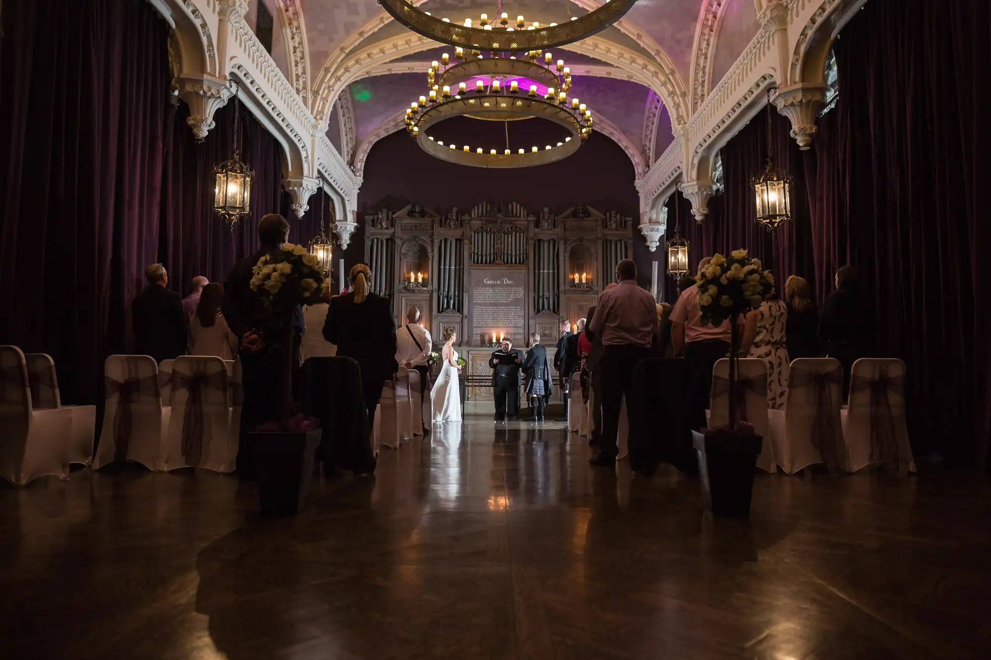 A wedding ceremony in an ornate hall, with guests standing as the couple walks down the aisle, floral decorations on either side.