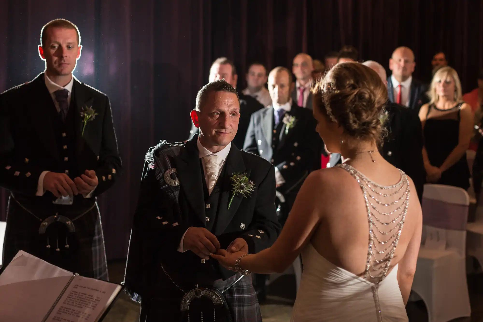 A bride and groom exchange rings at their wedding ceremony, surrounded by guests in a dimly lit room. the groom wears traditional scottish attire.