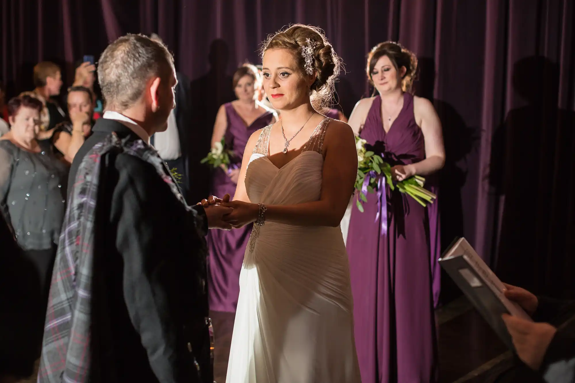 Bride in a strapless gown and groom in a tartan kilt exchange rings during a ceremony, with bridesmaids in purple dresses and guests in the background.
