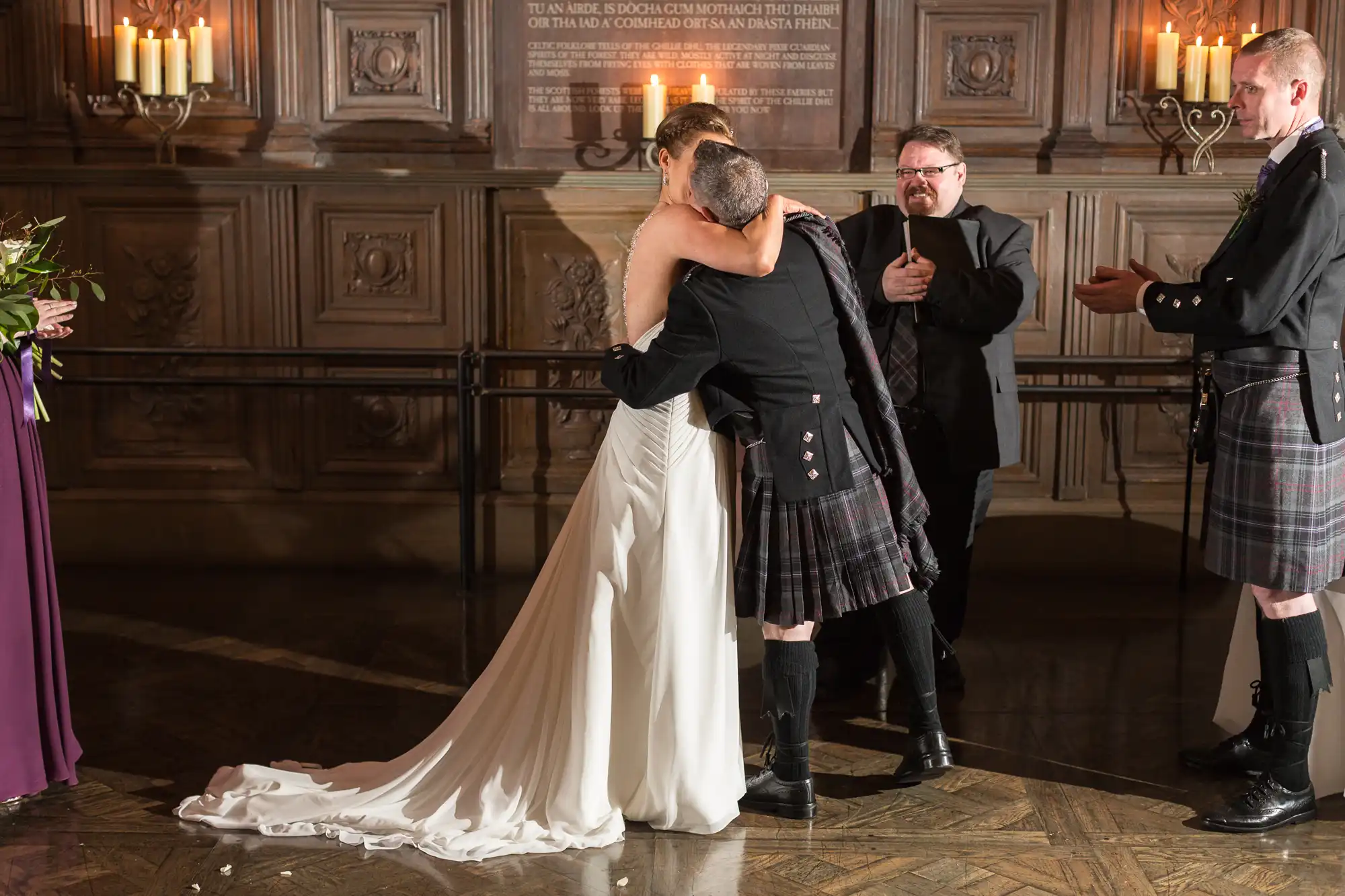 A bride and groom kissing, the groom wears a kilt. a man claps for them, and two people observe in a grand hall.