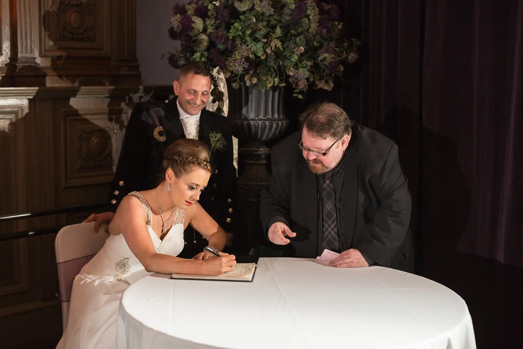 A bride and groom sign a document at a table, with a smiling officiant standing behind them in a dimly lit room decorated with flowers.