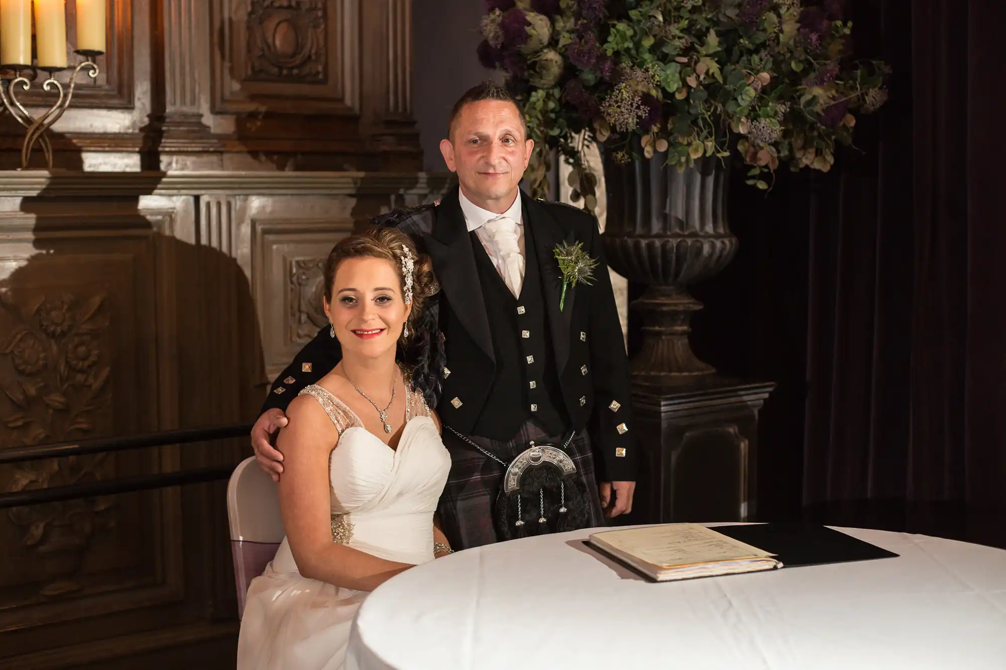 A bride and groom smiling, dressed in traditional scottish attire, standing beside a table in an opulent room.