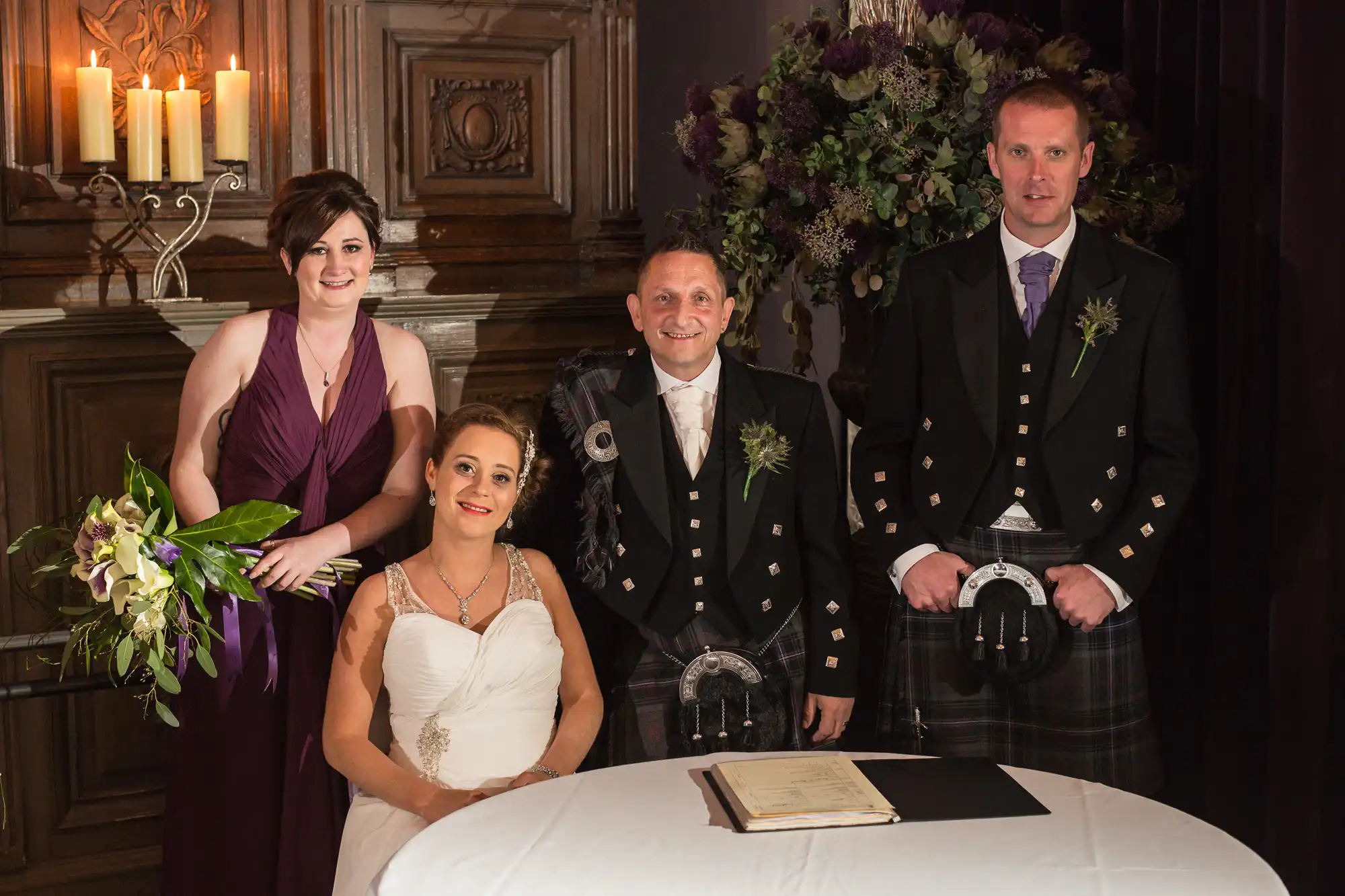 A wedding group with two men in kilts and two women, one in a bridal gown, standing by a table with a book, in a room with candles and floral decorations.