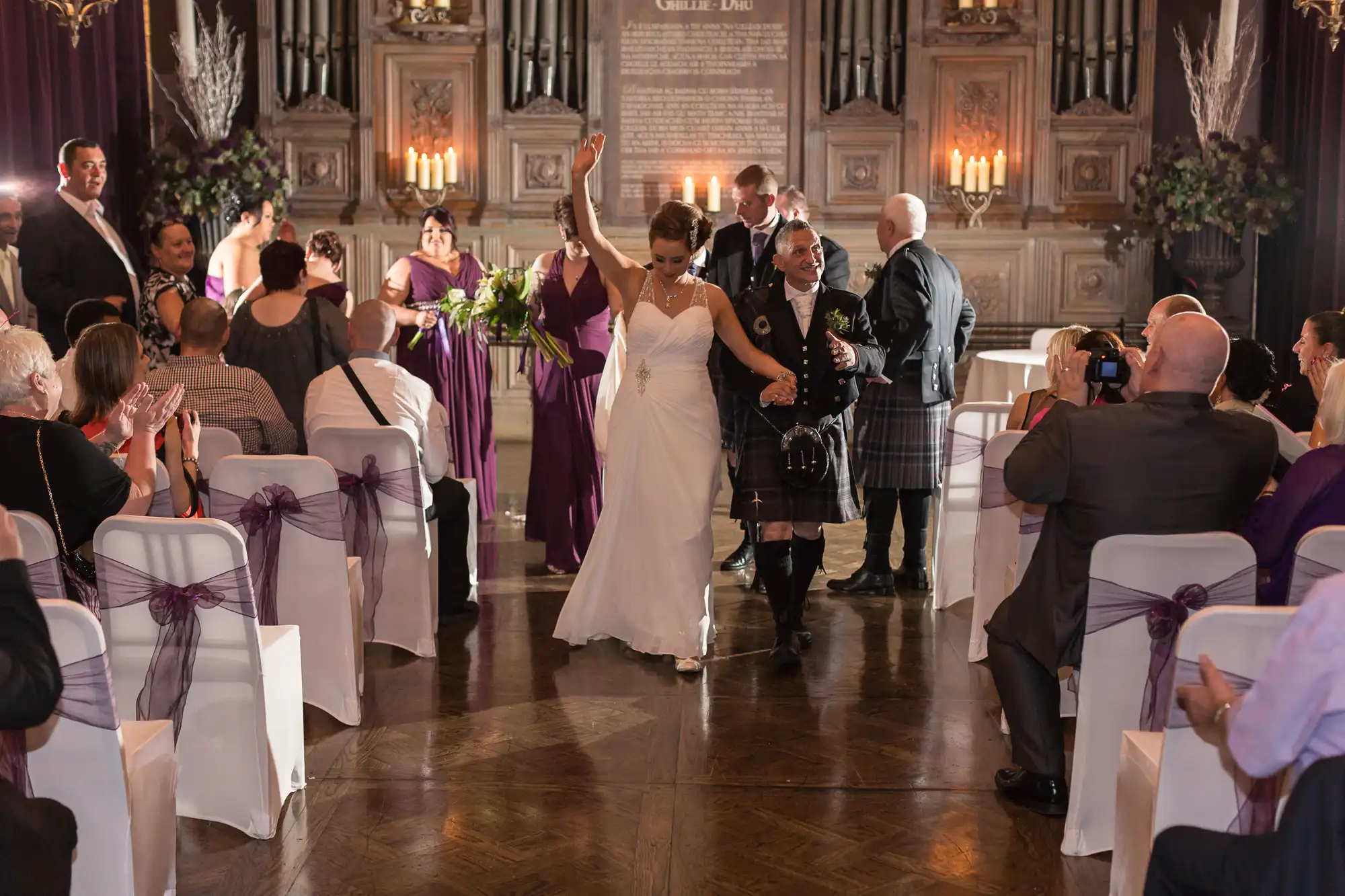 Bride and groom walking down the aisle, guests applauding, in a grand hall with elegant decor.