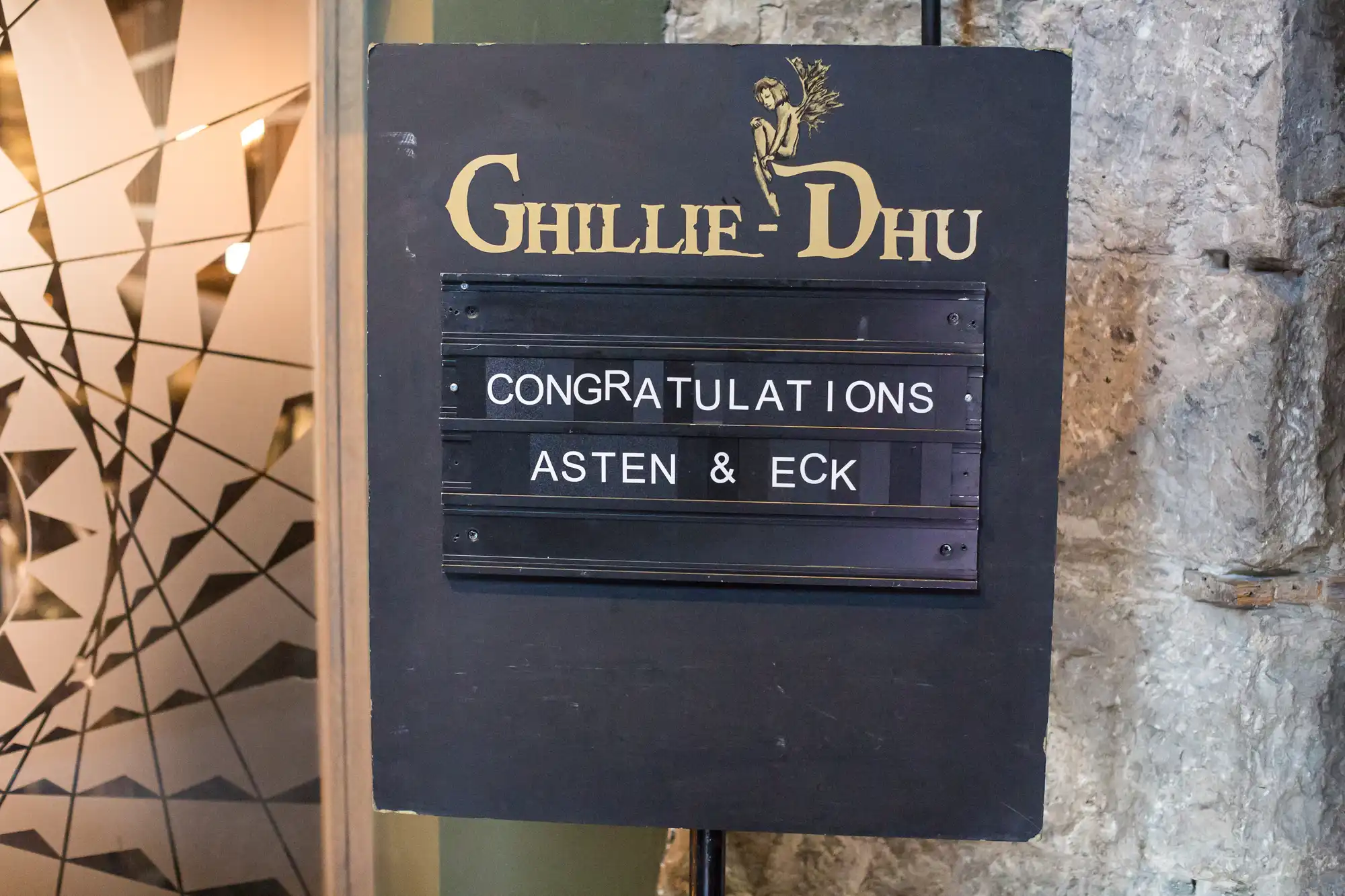 Signboard of ghillie-dhu pub displaying a message "congratulations asten & eck" in front of a textured wall.