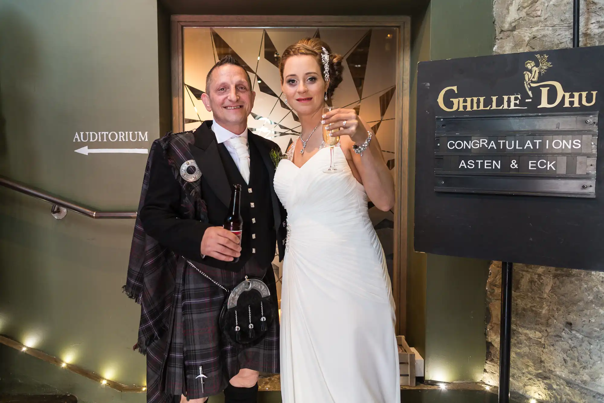 A newlywed couple in formal attire, standing beside a sign that reads "congratulations asten & eck" at ghillie dhu.