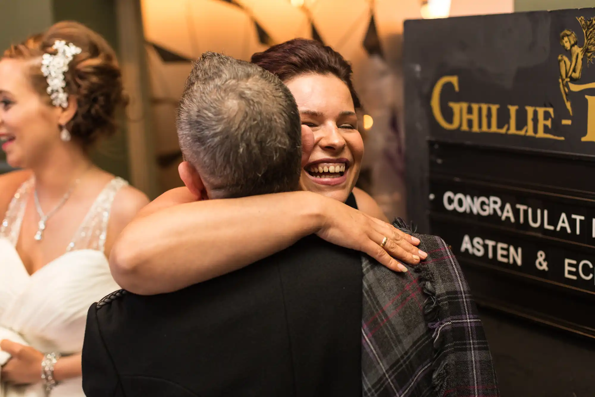 A joyful bride hugs a man in a kilt at a wedding reception, both smiling happily near a congratulatory sign.