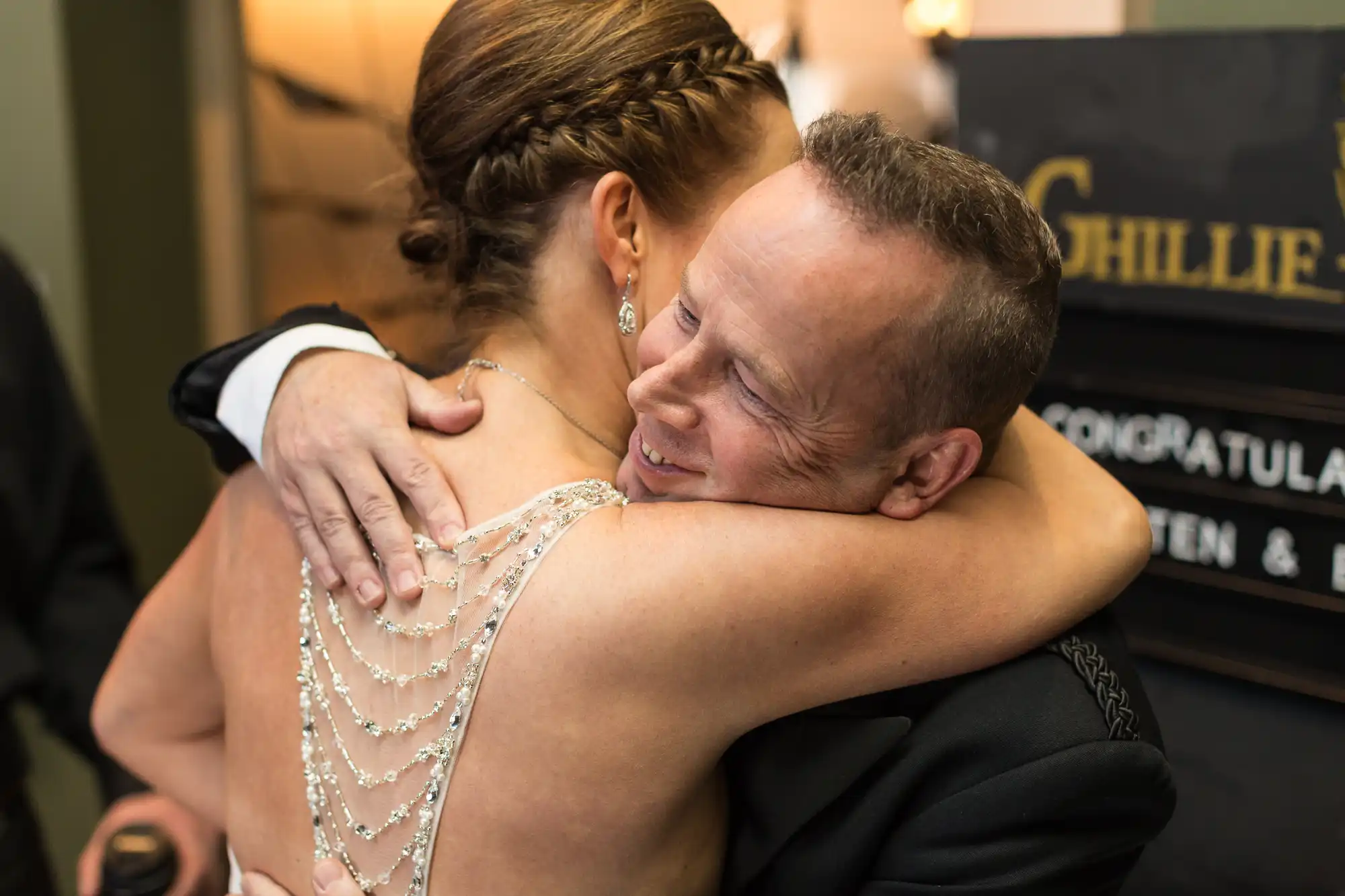 A man in a tuxedo embracing a woman in a beaded dress, both smiling joyfully, at an indoor celebration.