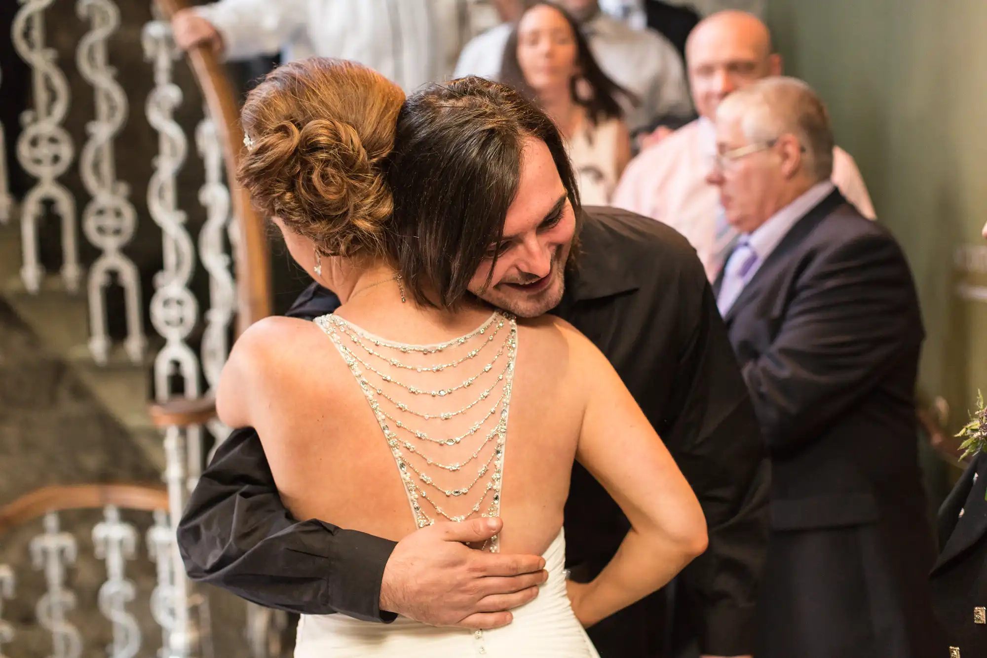 Bride in a white dress and groom embracing at their wedding ceremony, both smiling with guests in the background.