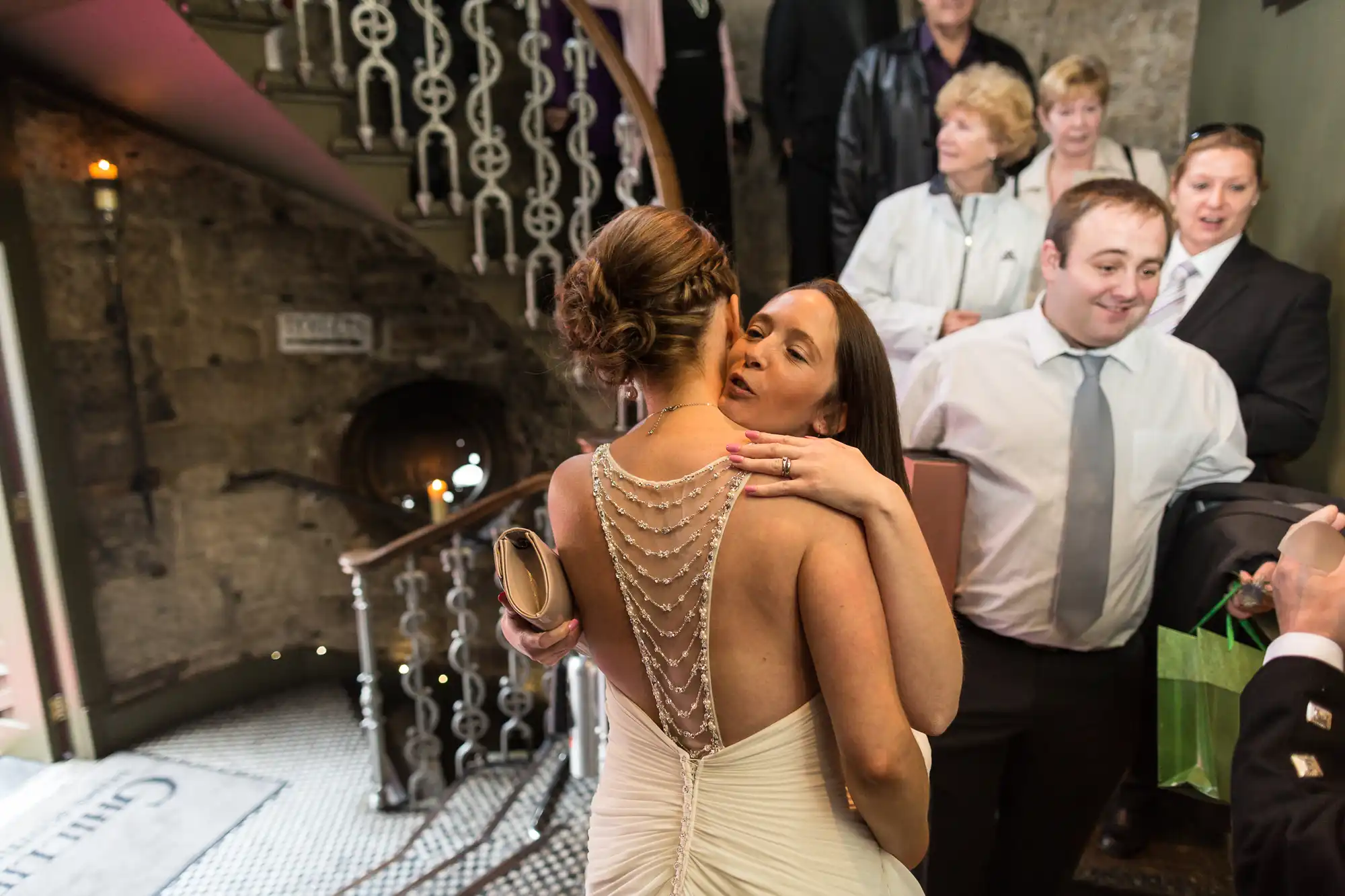A bride in a backless dress embraces a woman at a wedding, surrounded by guests watching and smiling.