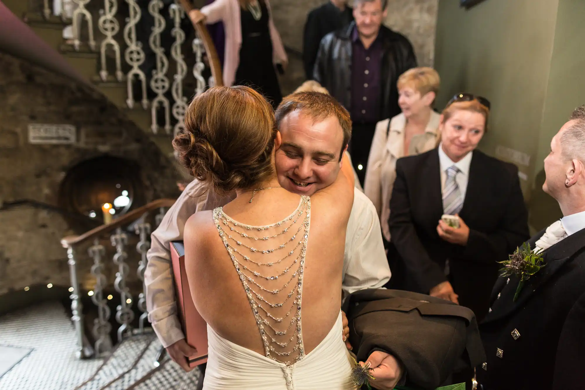 A bride and groom embrace warmly at a wedding reception, guests smiling in the background near a staircase with decorative railings.