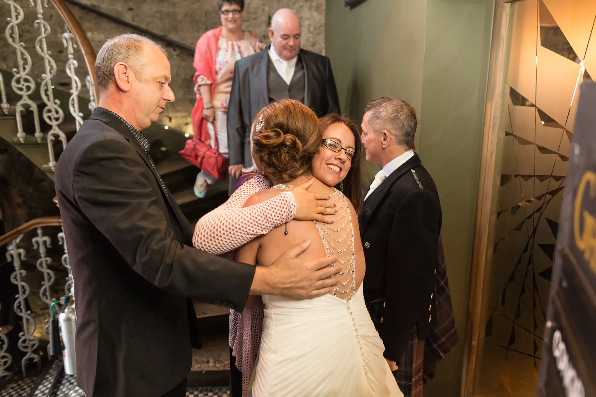 A bride in a white dress embracing a woman at the bottom of a staircase, with three guests smiling and looking on.