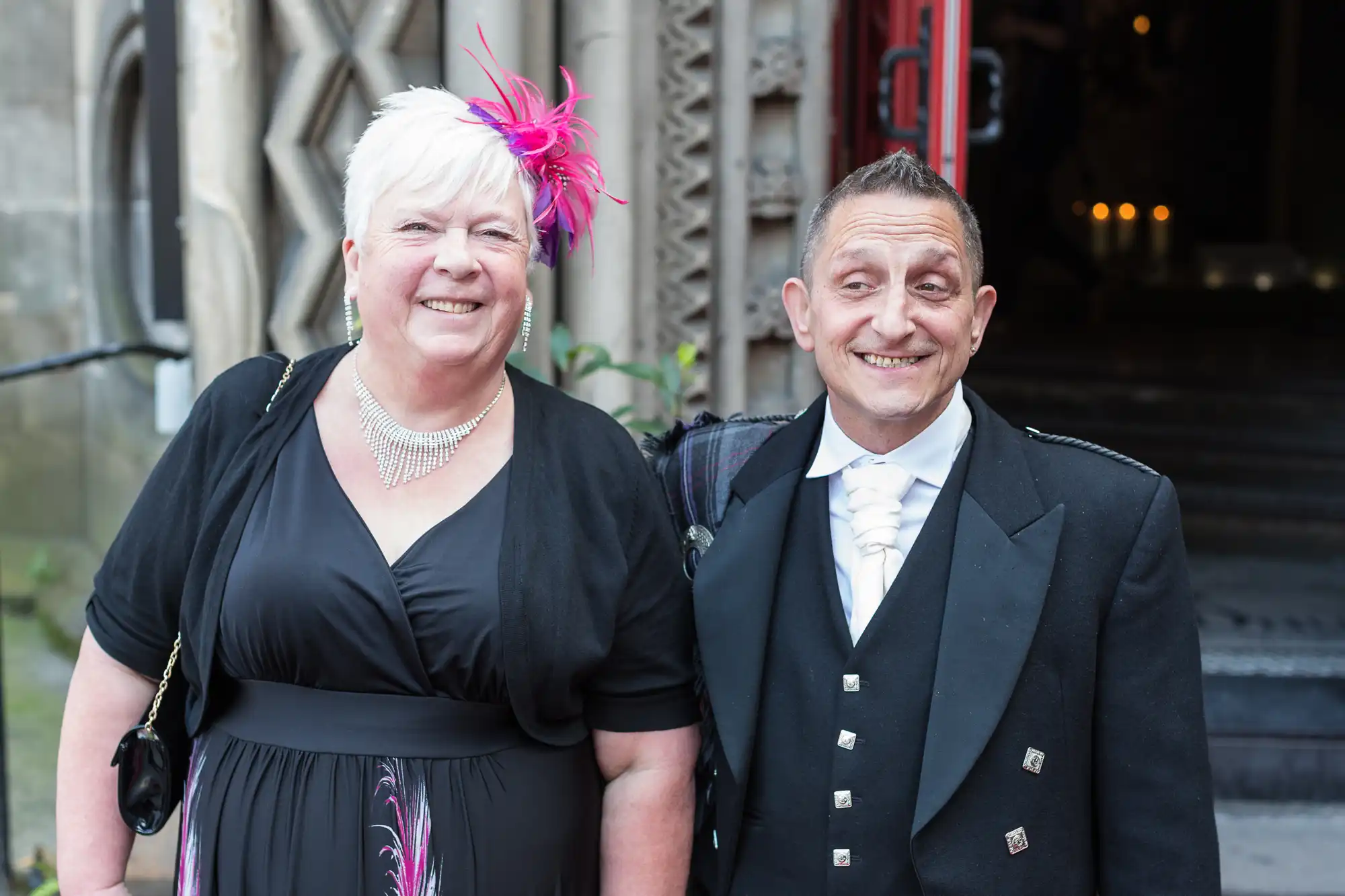 A smiling elderly couple dressed in formal attire standing outside a building, the woman wearing a black dress and pink fascinator, and the man in a black suit with medal decorations.