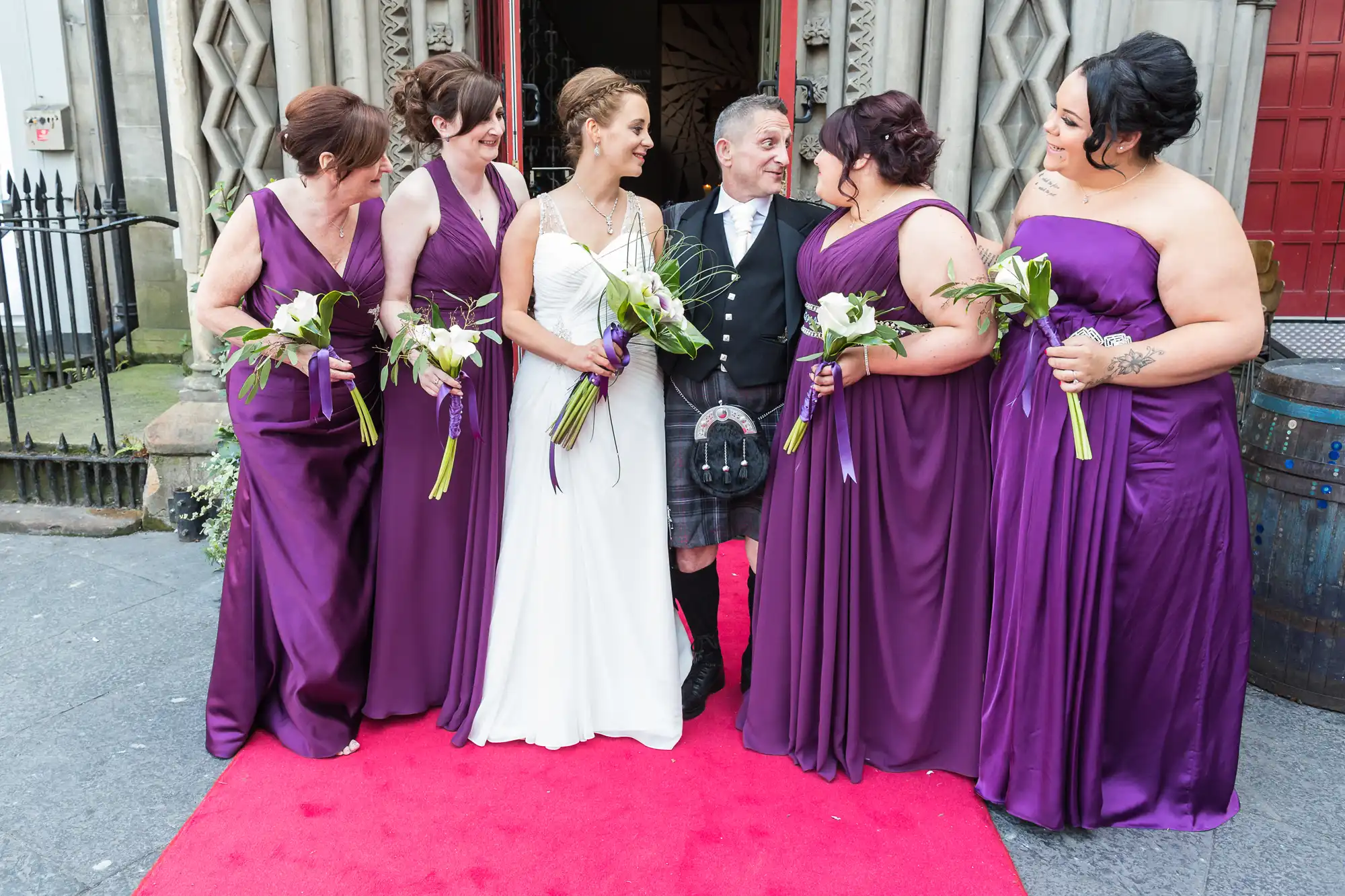 A bride and groom smile and chat with four bridesmaids dressed in purple on a red carpet outside a venue.