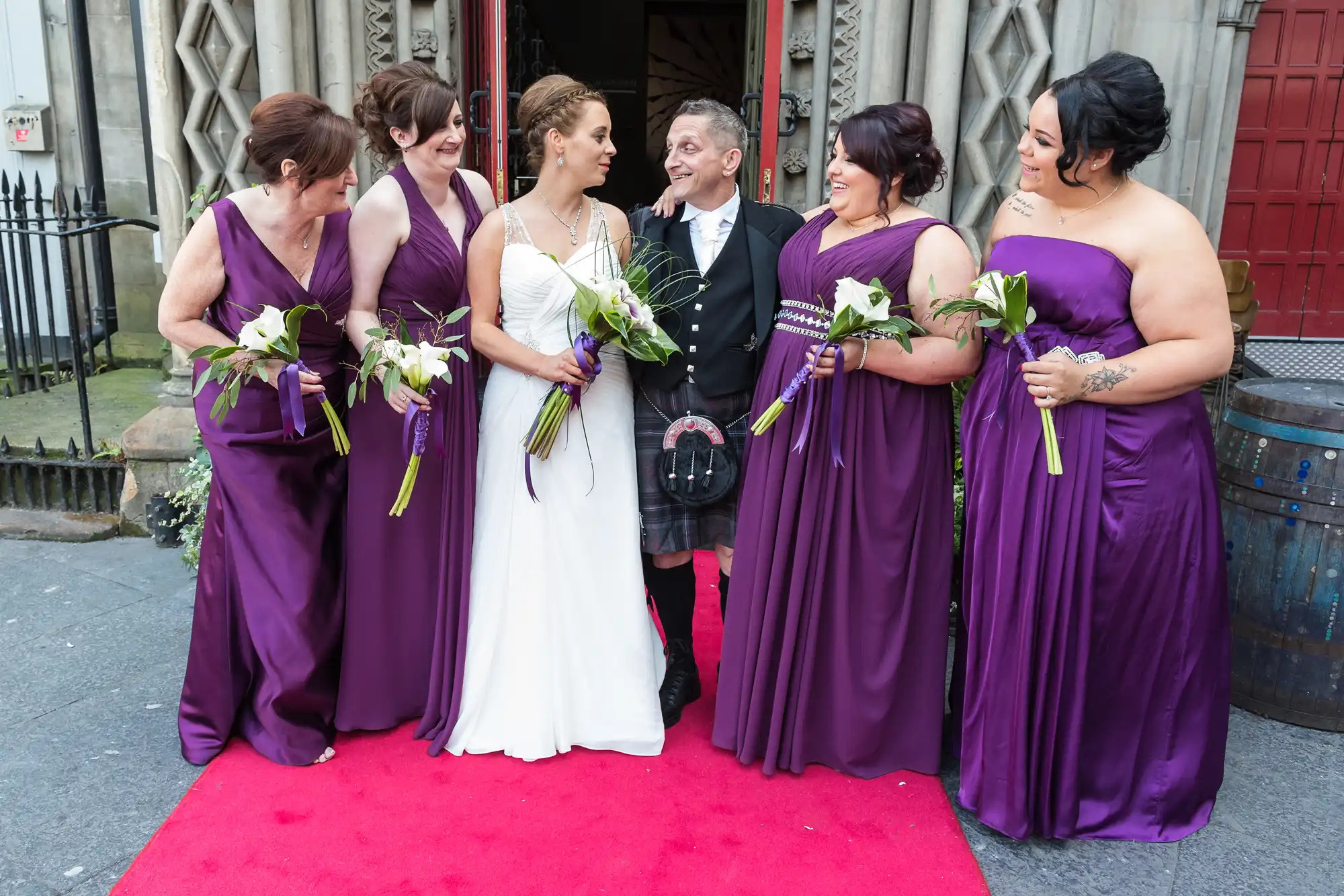 A bride and groom smiling and interacting joyfully with four bridesmaids in purple dresses, holding bouquets, on a red carpet outside a building.