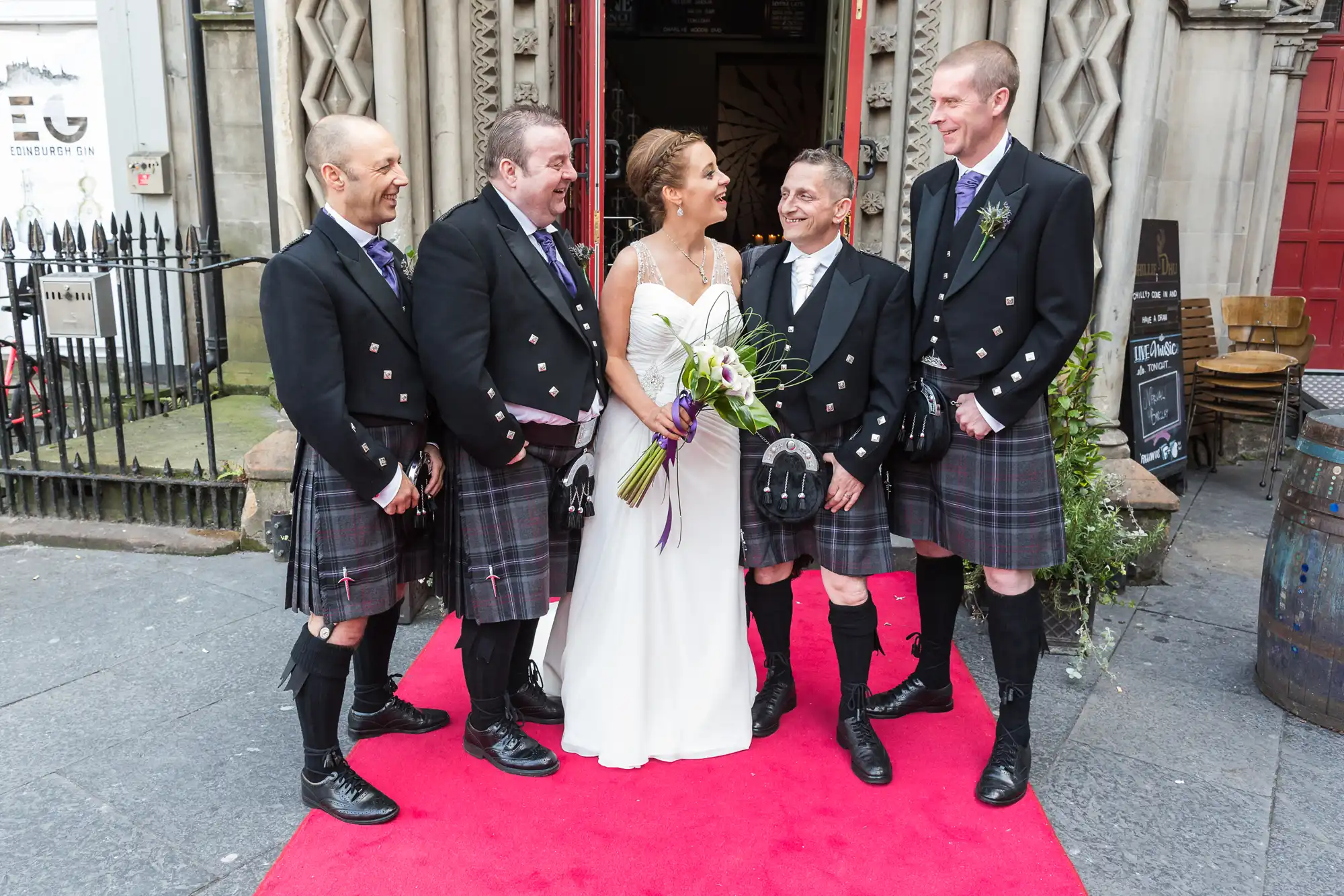 A bride and groom laugh with three groomsmen in kilts outside a building with a red carpet entrance.
