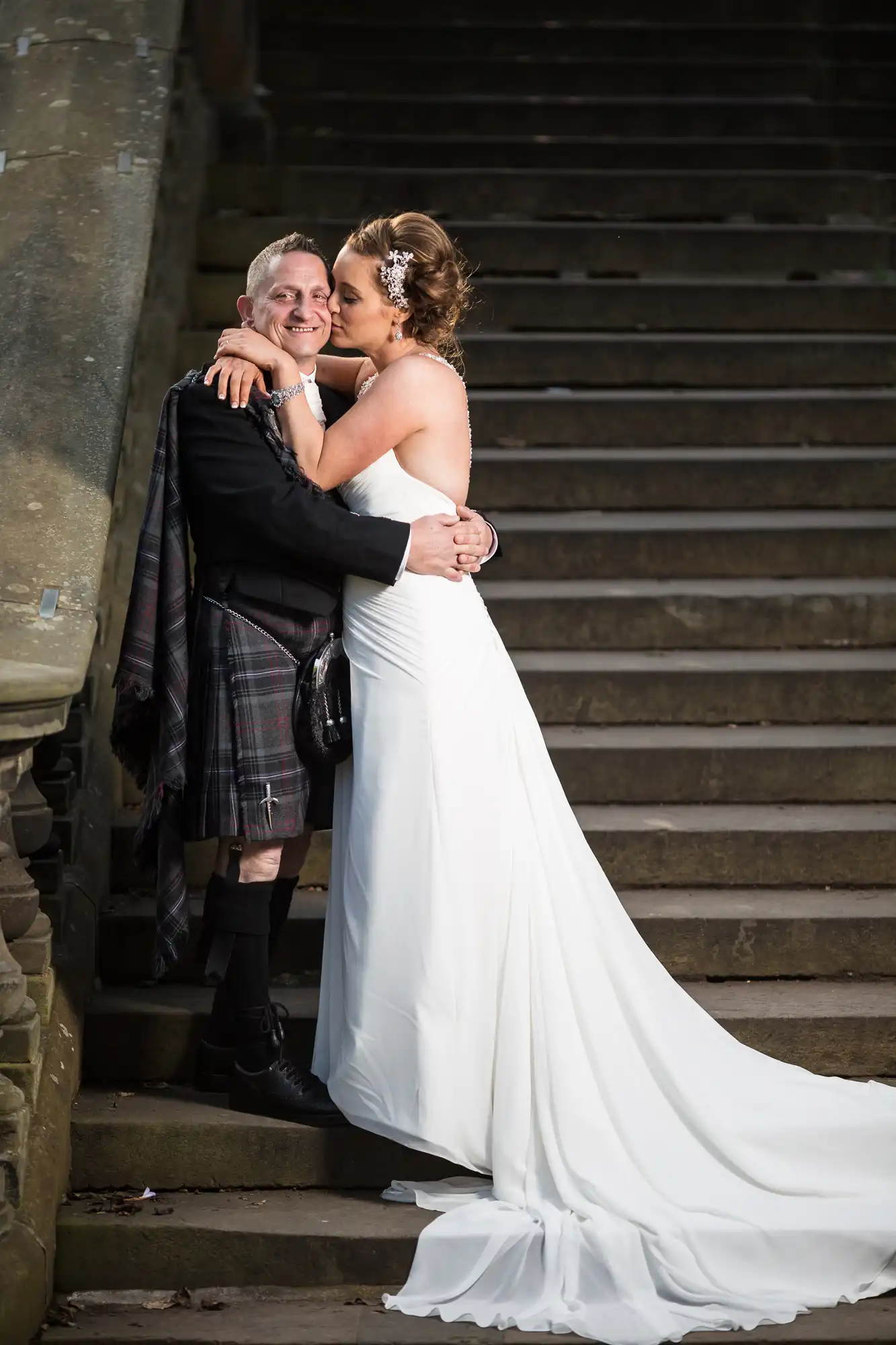 A newly married couple embracing on stone stairs, the groom in a kilt and the bride in a white gown with a long train.
