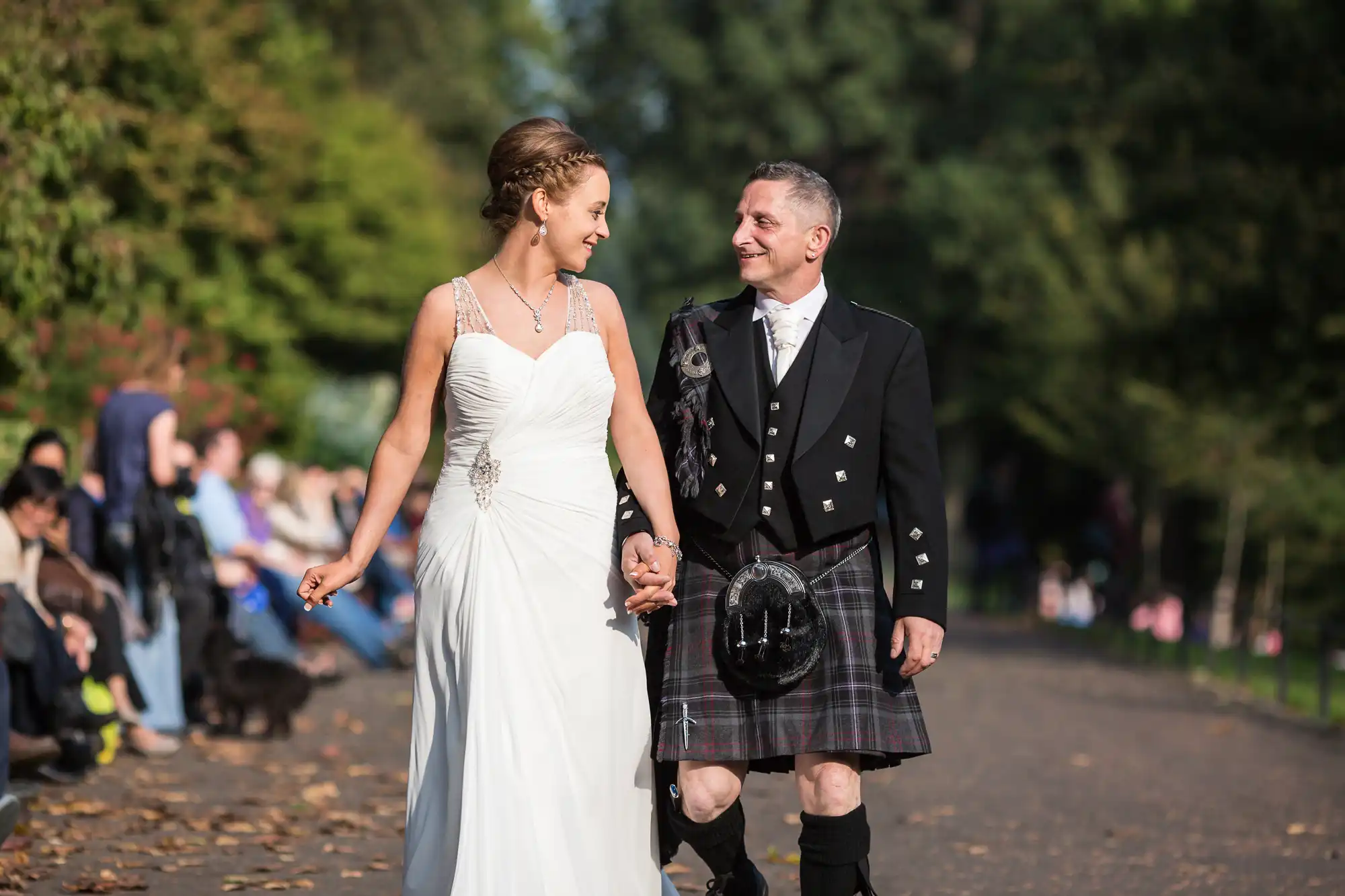 Bride in a white dress and groom in a kilt smiling and holding hands while walking down a park alley, with spectators seated on benches.
